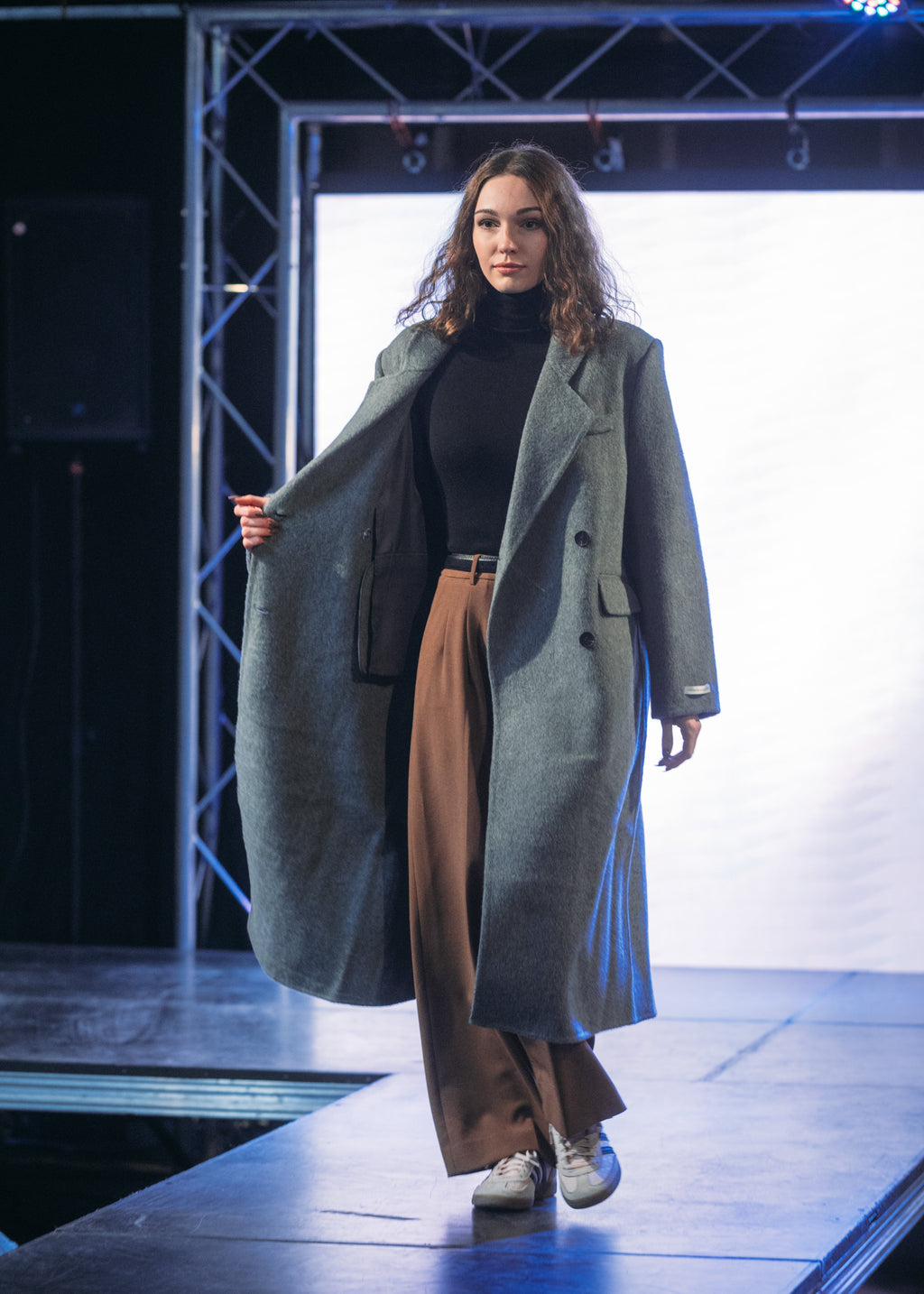 A model walks down a runway wearing a long charcoal gray double-breasted overcoat over a black turtleneck, brown wide-leg trousers, and white sneakers.