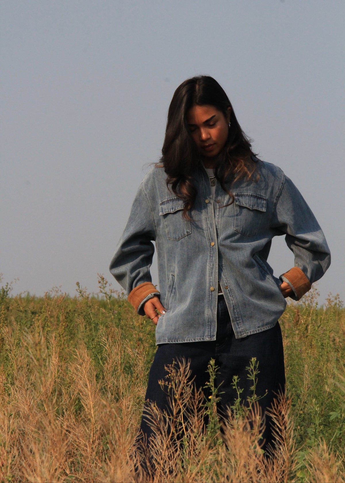 A woman standing in a grassy field wearing a light blue denim jacket with brown cuffs and dark pants, looking down.