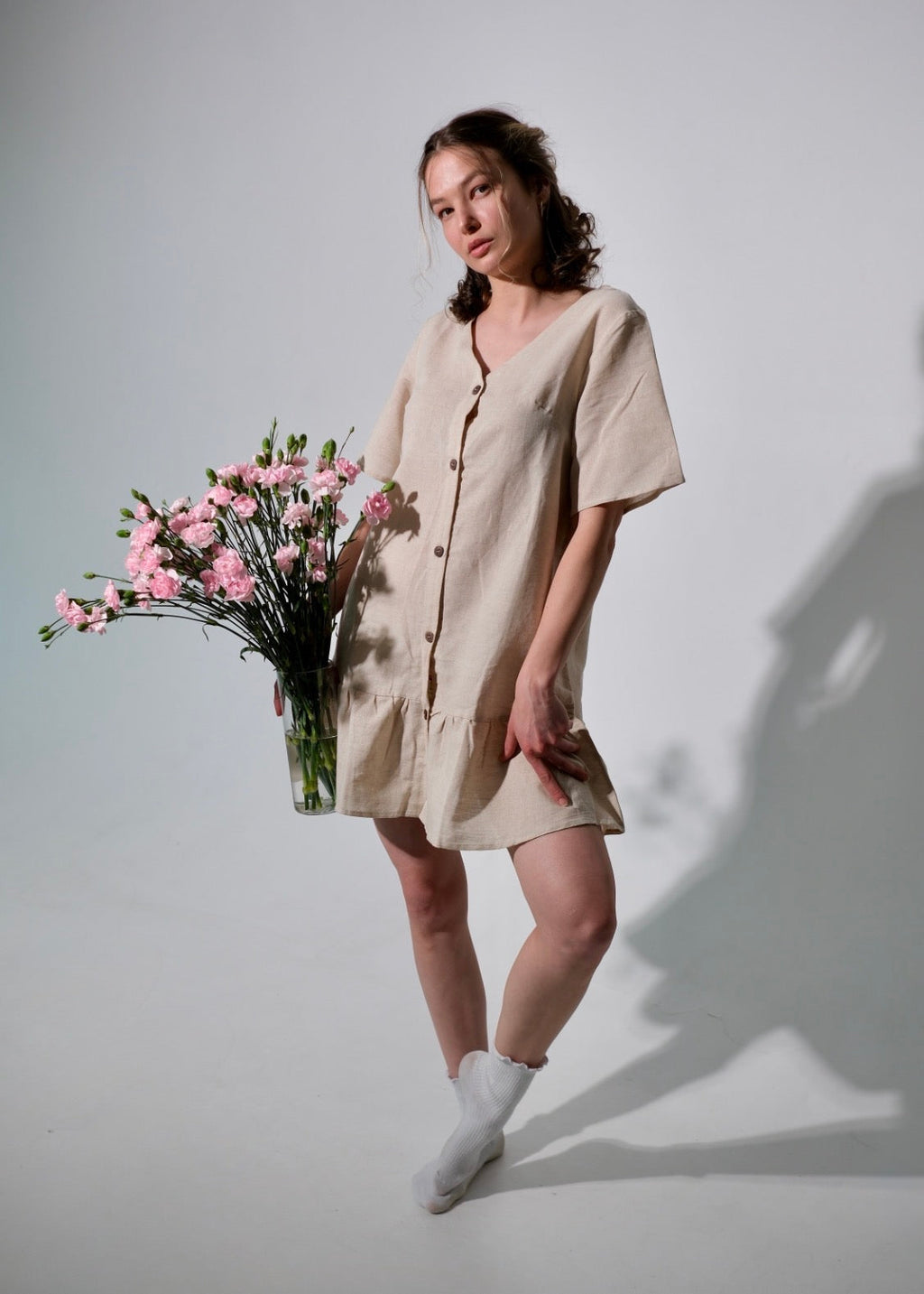 Woman in a beige dress holding pink flowers against a plain background