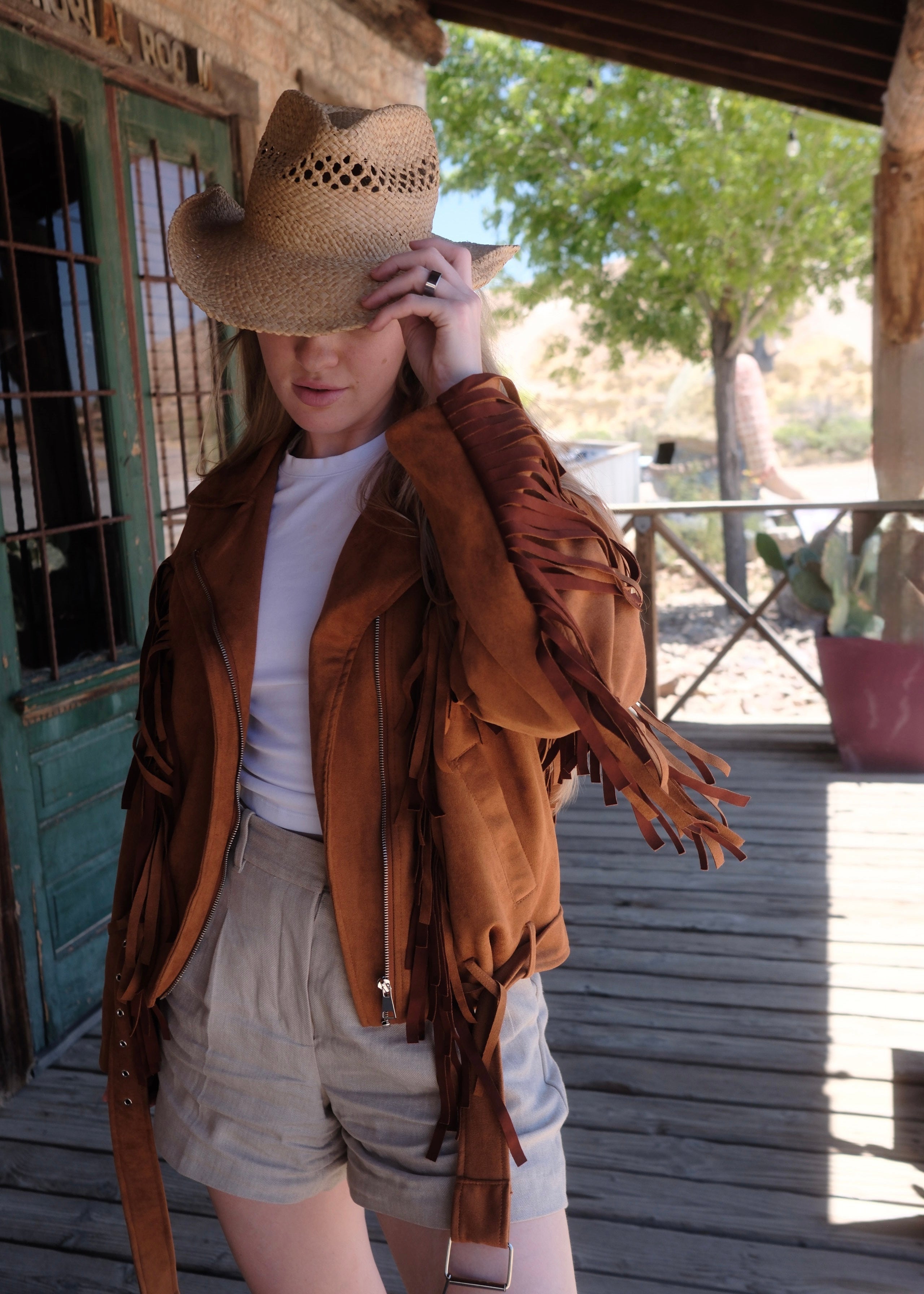 Person wearing a brown leather jacket and straw hat in front of a rustic building.