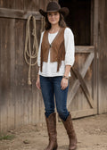 Woman wearing a cowboy hat, fringed vest, white blouse, blue jeans, and brown boots in front of a wooden barn.