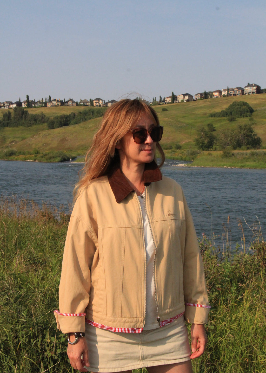 A woman wearing a beige zip-up jacket with brown collar and pink trim, a white top, and a beige mini skirt, standing outdoors in front of a lake with grassy hills and houses in the background.