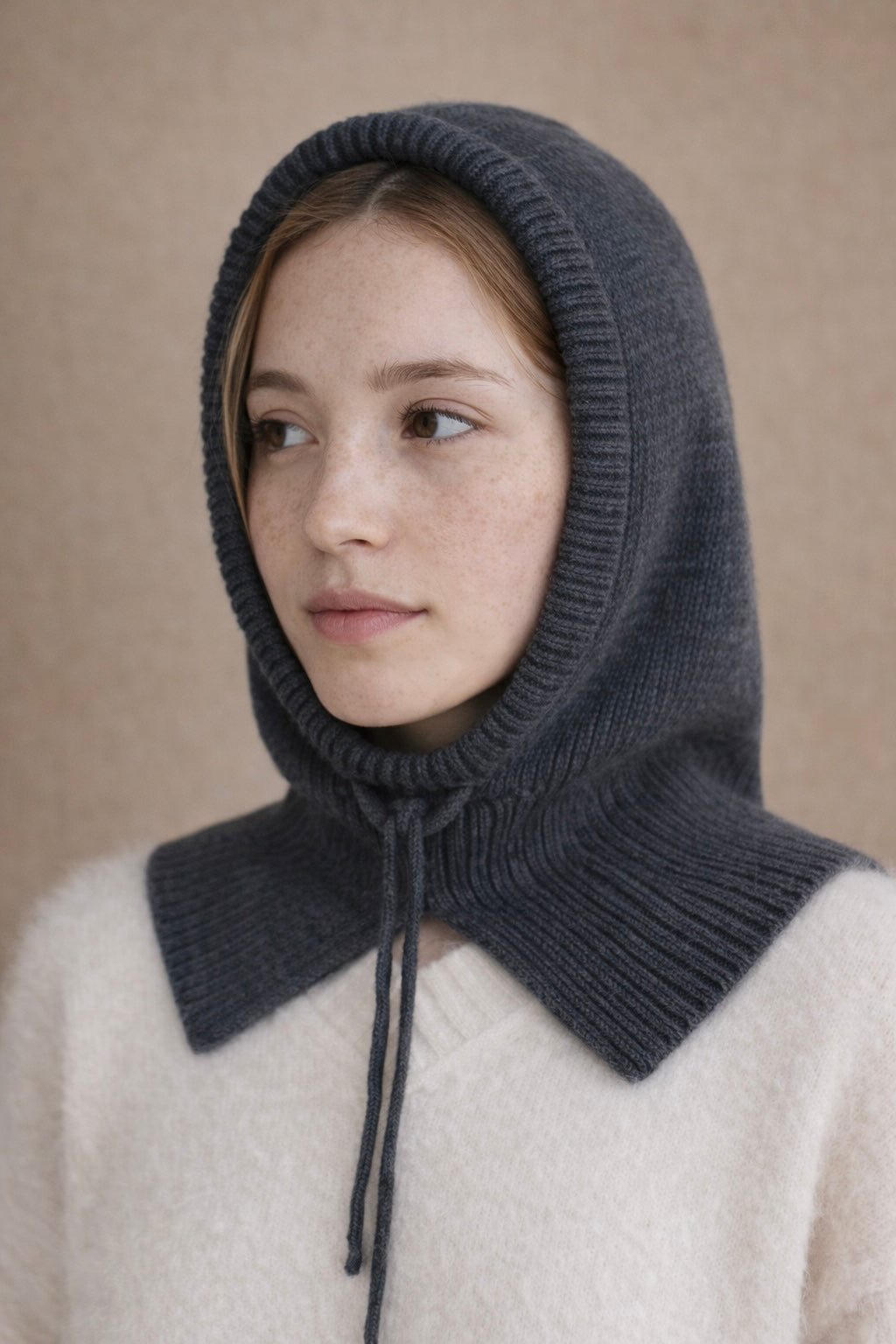 Person wearing a dark gray knitted hood over a light-colored sweater against a beige background