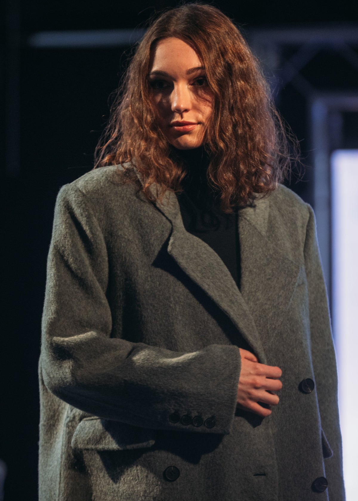 A woman with long, wavy brown hair wearing a textured charcoal gray double-breasted coat over a black top, captured in a dimly lit indoor setting.