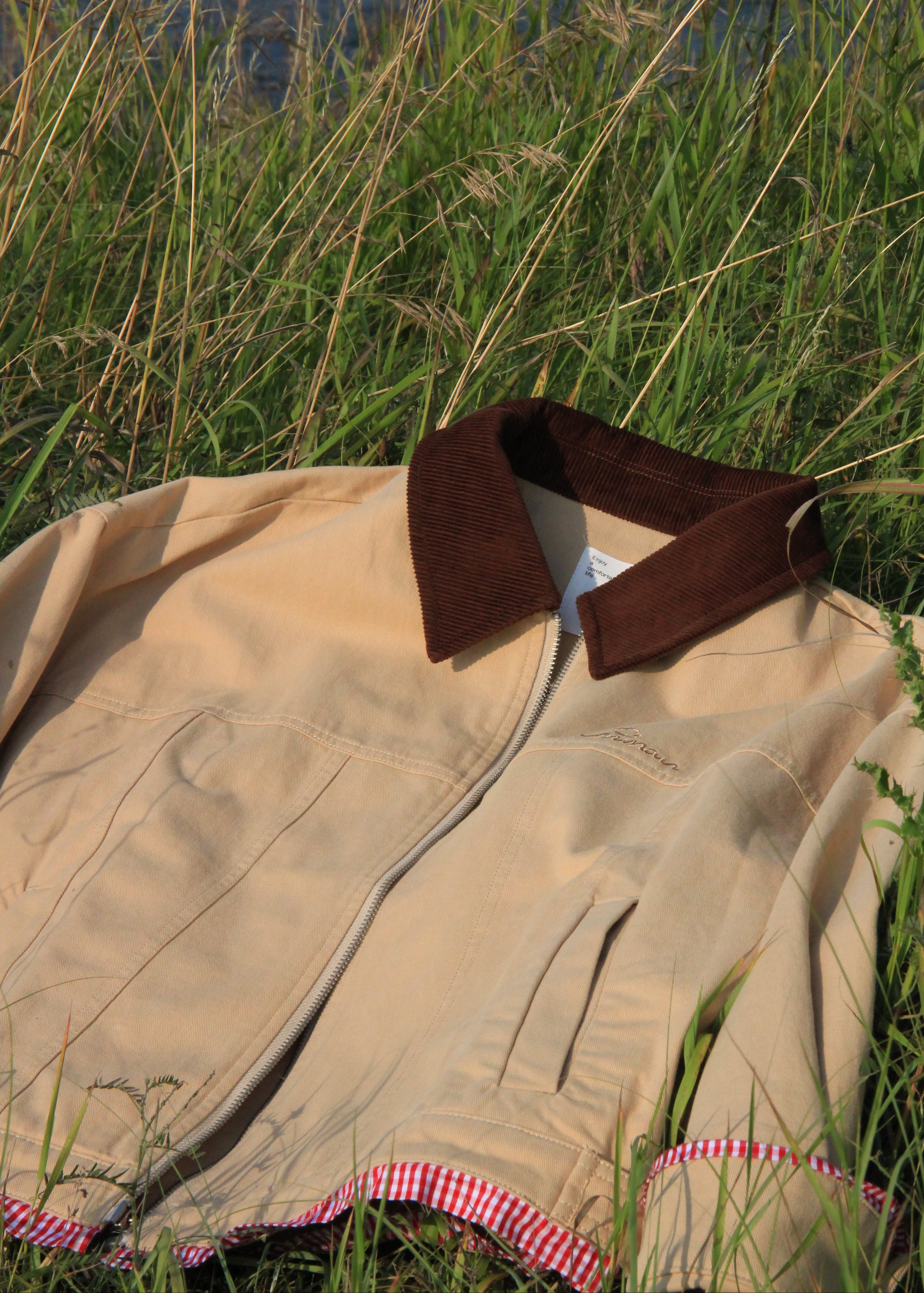 A beige jacket laid on tall green grass near water, featuring a dark brown corduroy collar and a red-and-white gingham hem.
