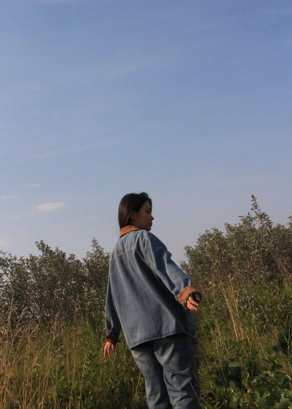 A person standing outdoors in a grassy field under a clear blue sky, wearing a light blue denim jacket and matching blue jeans.