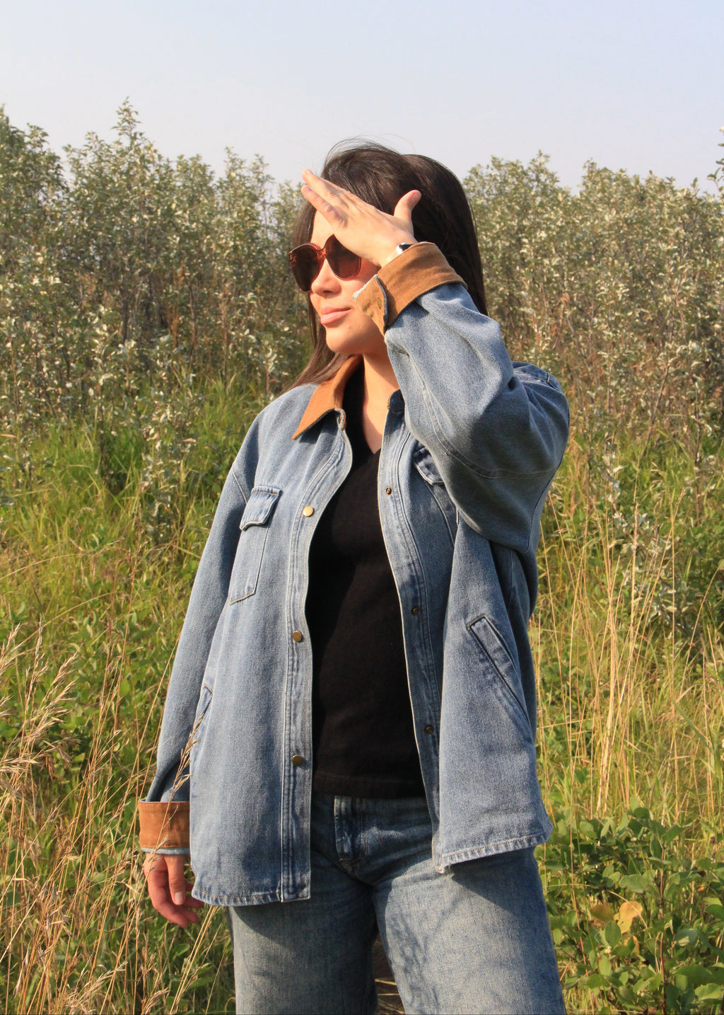 A woman wearing a light blue denim jacket with brown collar and cuff trim, a black top, and blue jeans, standing outdoors in a grassy field and shielding her eyes from the sun.
