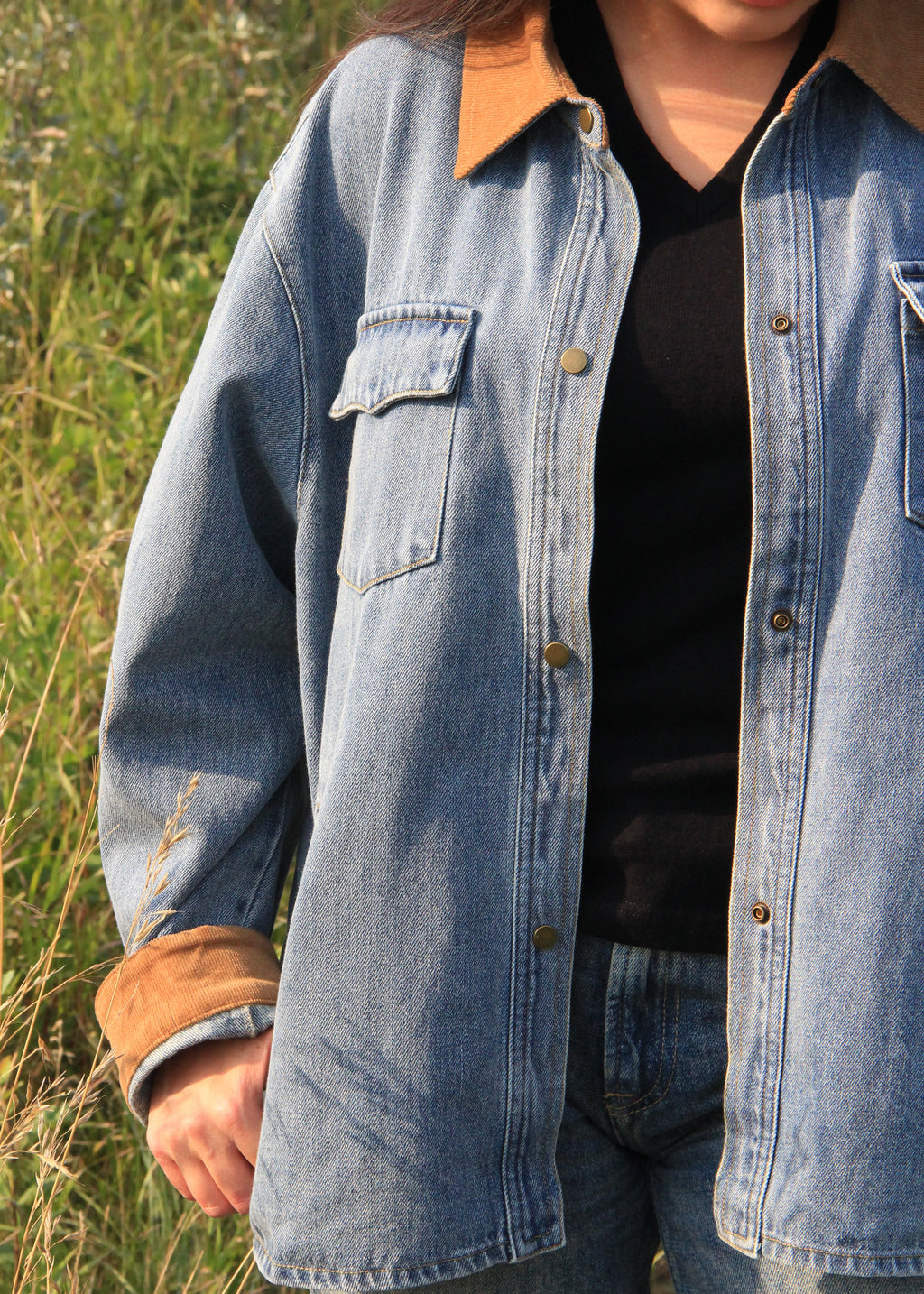 A person wearing an oversized light blue denim shirt jacket with a tan collar and turned-up cuffs over a black top, photographed outdoors in a grassy field.
