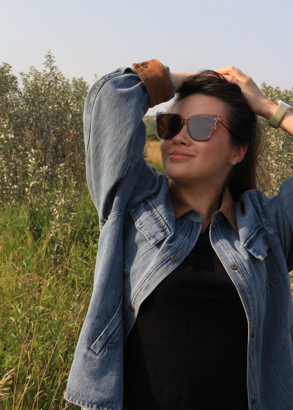 A woman outdoors in a grassy field wearing a light blue denim jacket over a black top, with brown sunglasses and her hands in her hair.