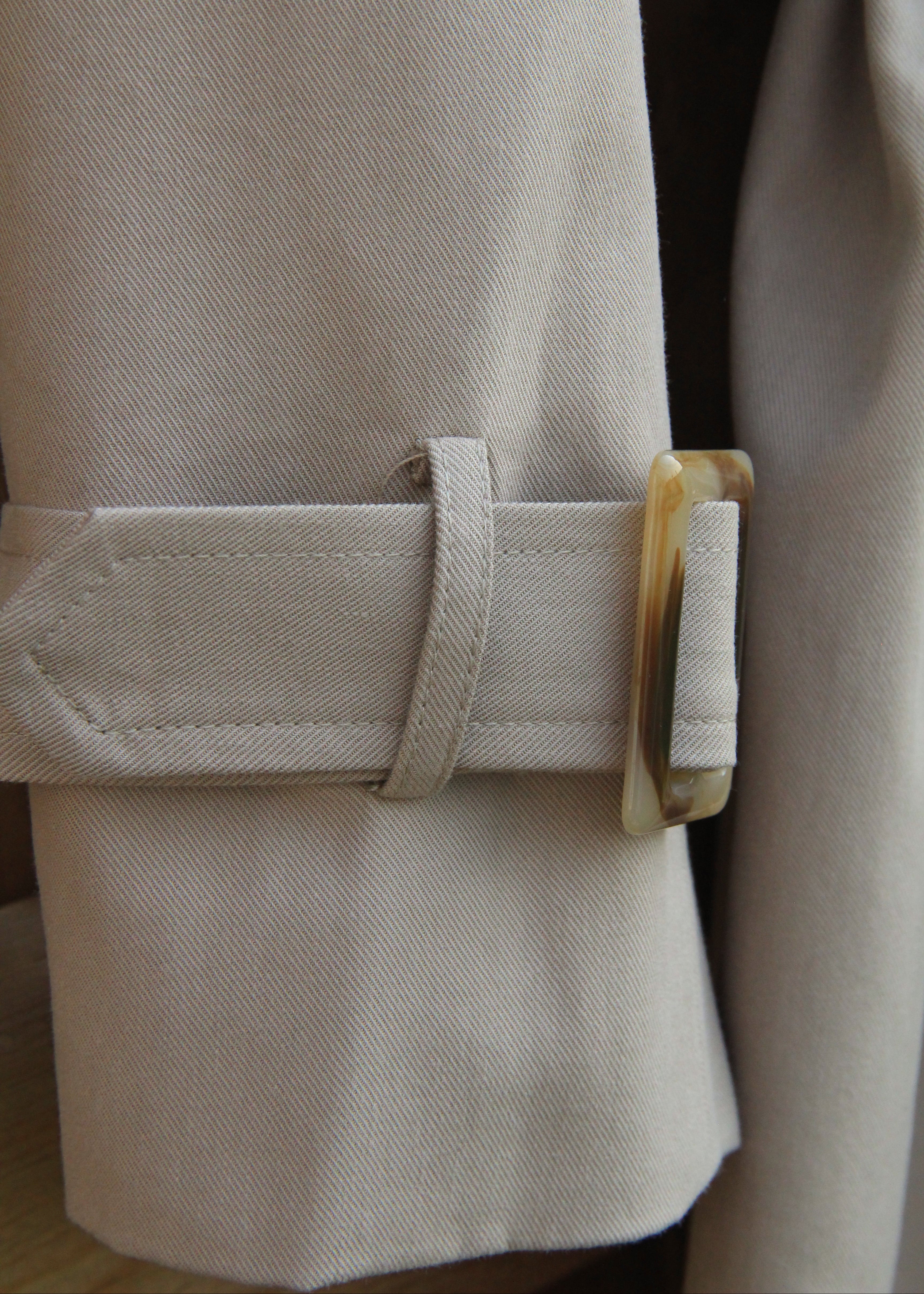 Close-up of a light beige trench coat sleeve with a matching fabric cuff strap and a decorative amber-brown rectangular buckle.
