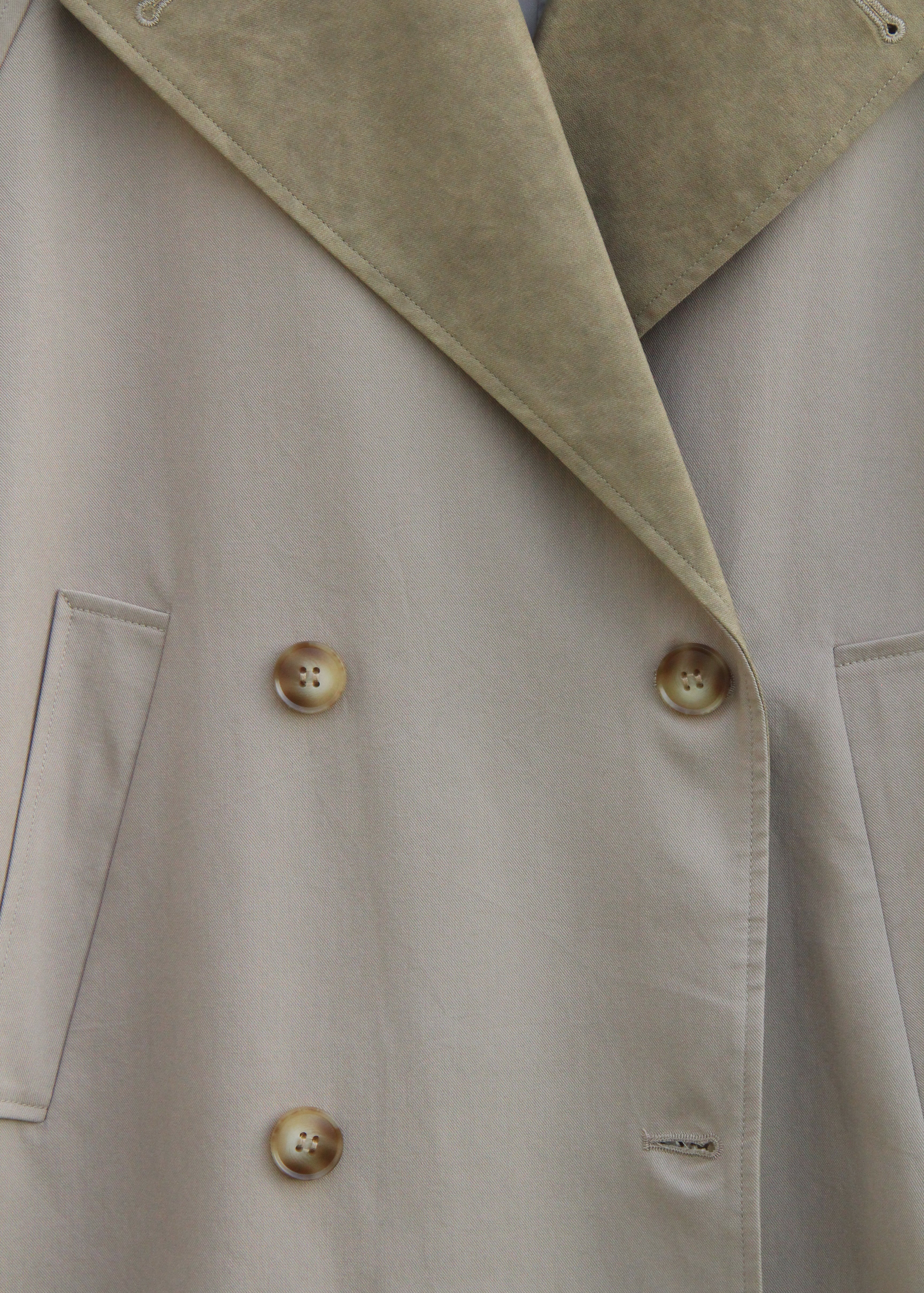 Close-up of a double-breasted coat with a light beige body and a contrasting tan suede-like lapel, featuring decorative buttons.