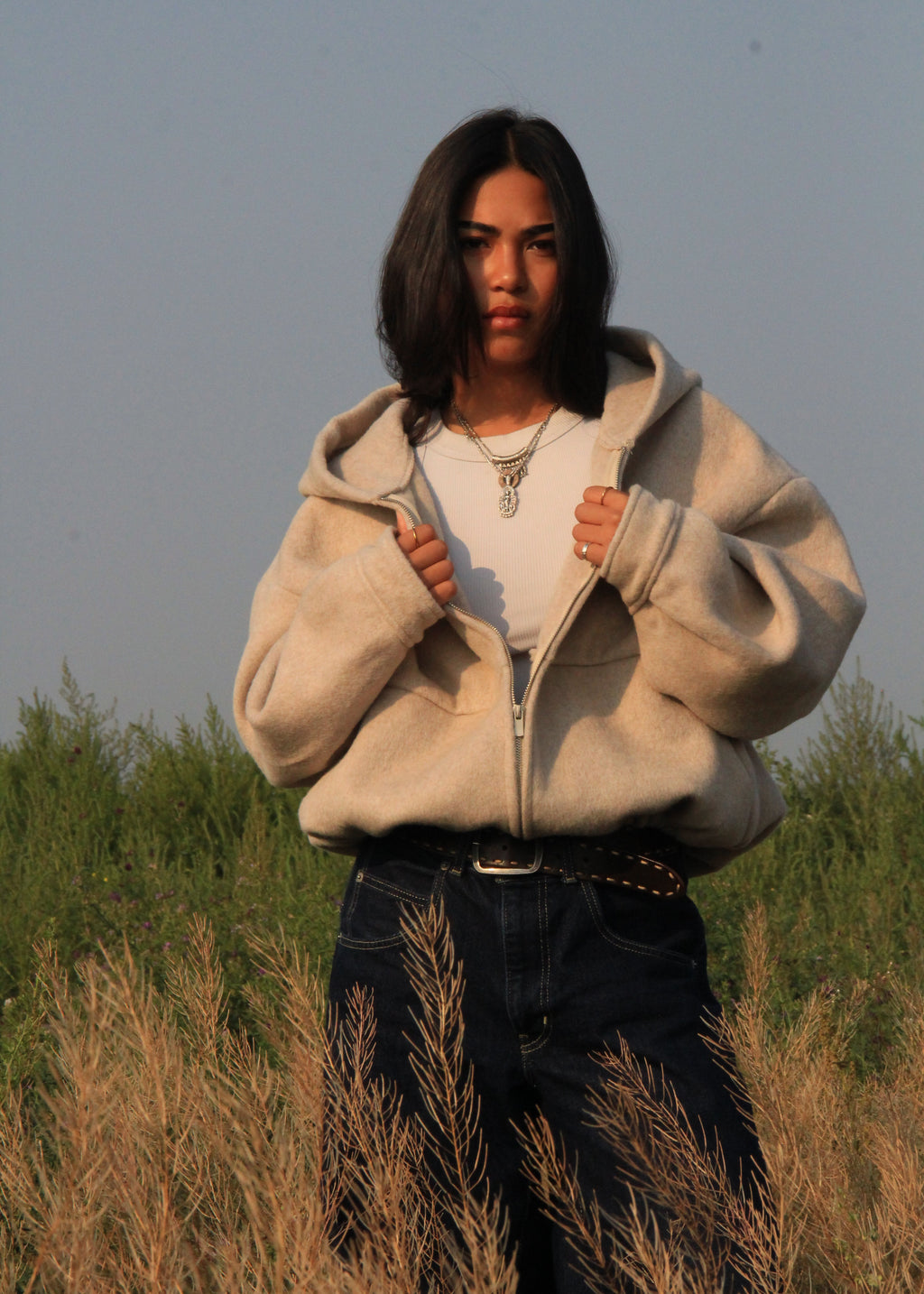 A woman stands in a field wearing a cream-colored zip-up fleece hoodie over a white t-shirt and dark blue jeans.