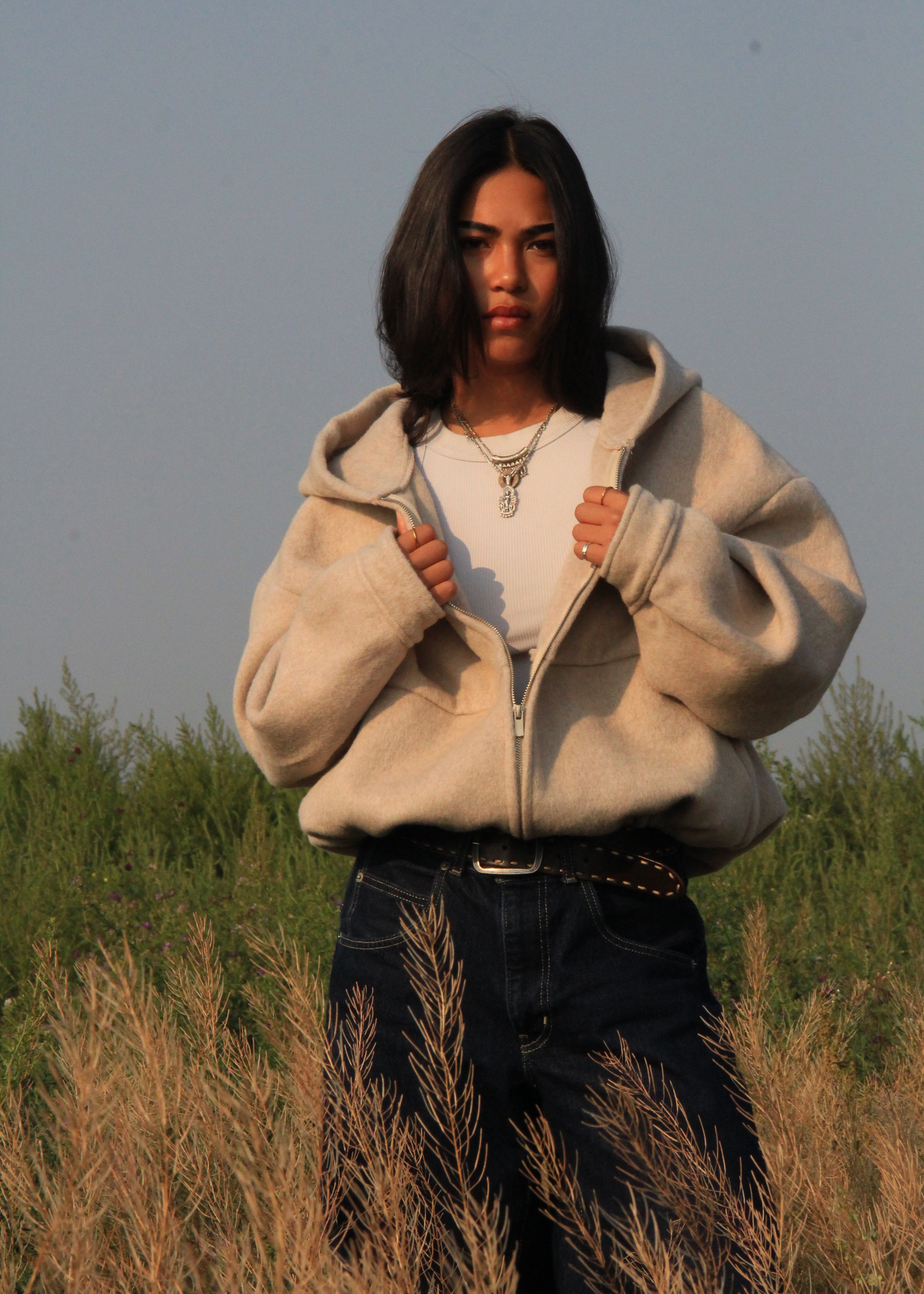 A woman stands in a field wearing a cream-colored zip-up fleece hoodie over a white t-shirt and dark blue jeans.