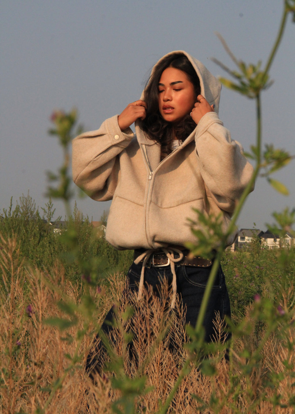 A woman wearing a beige sherpa zip-up hoodie and dark jeans stands in a grassy field at sunset, with blurred green plants in the foreground.