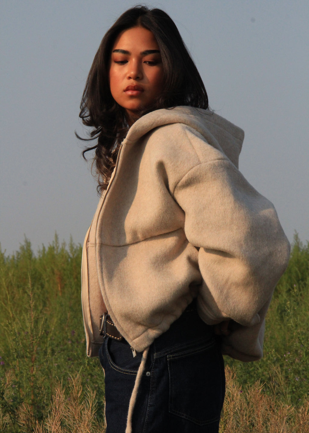 A woman wearing a beige oversized cropped hoodie and dark blue jeans, standing outdoors in a grassy field under a clear sky.