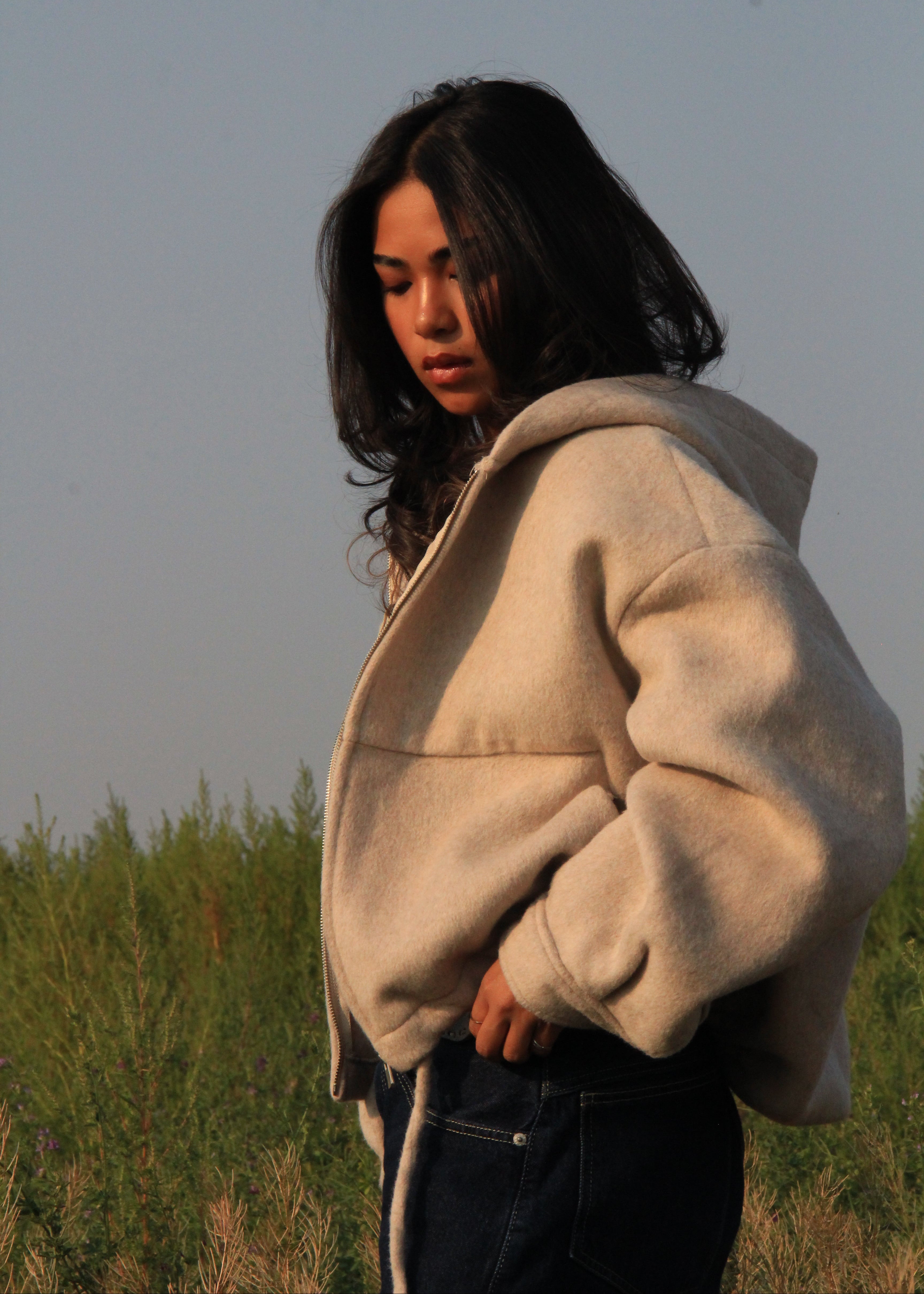 A woman standing outdoors in a grassy field at golden hour, wearing a cream-colored oversized fleece hoodie and dark blue jeans.