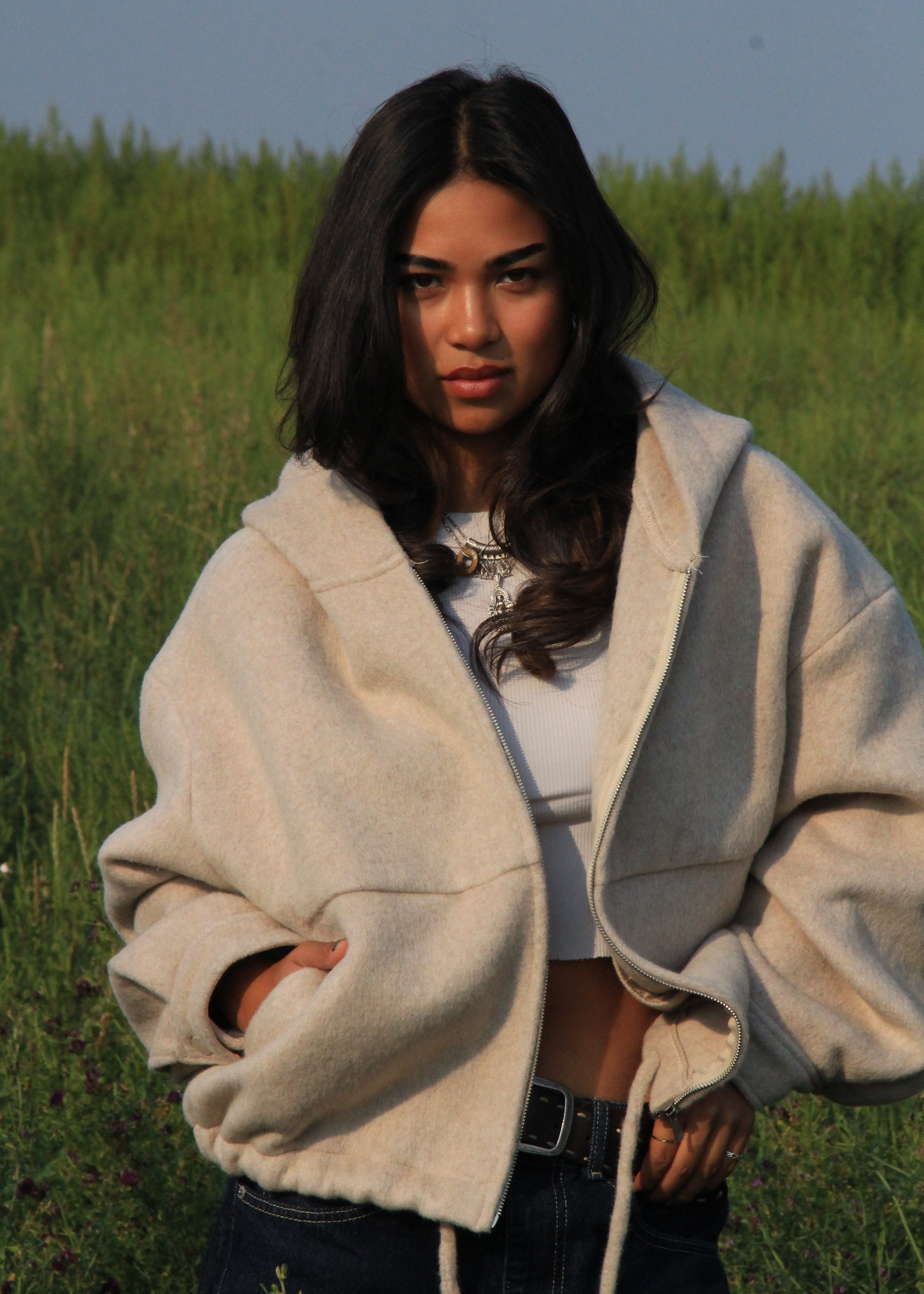 A woman wearing a cream-colored oversized zip-up hoodie with drawstrings over a white crop top and dark jeans, standing in a grassy field.