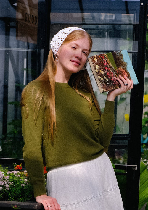 Woman in a green sweater and white skirt holding a book in a greenhouse setting.