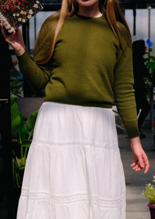 Person wearing a green sweater and white skirt outdoors with plants in the background