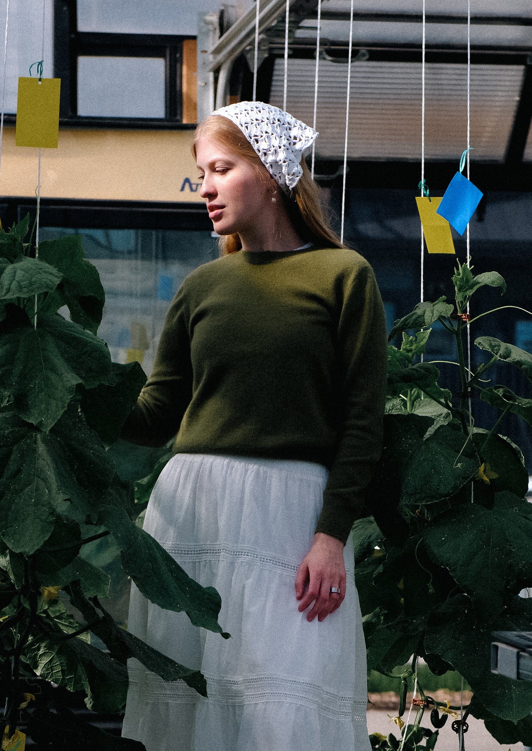 Woman in a green sweater and white skirt standing in a greenhouse with plants and tables around her.