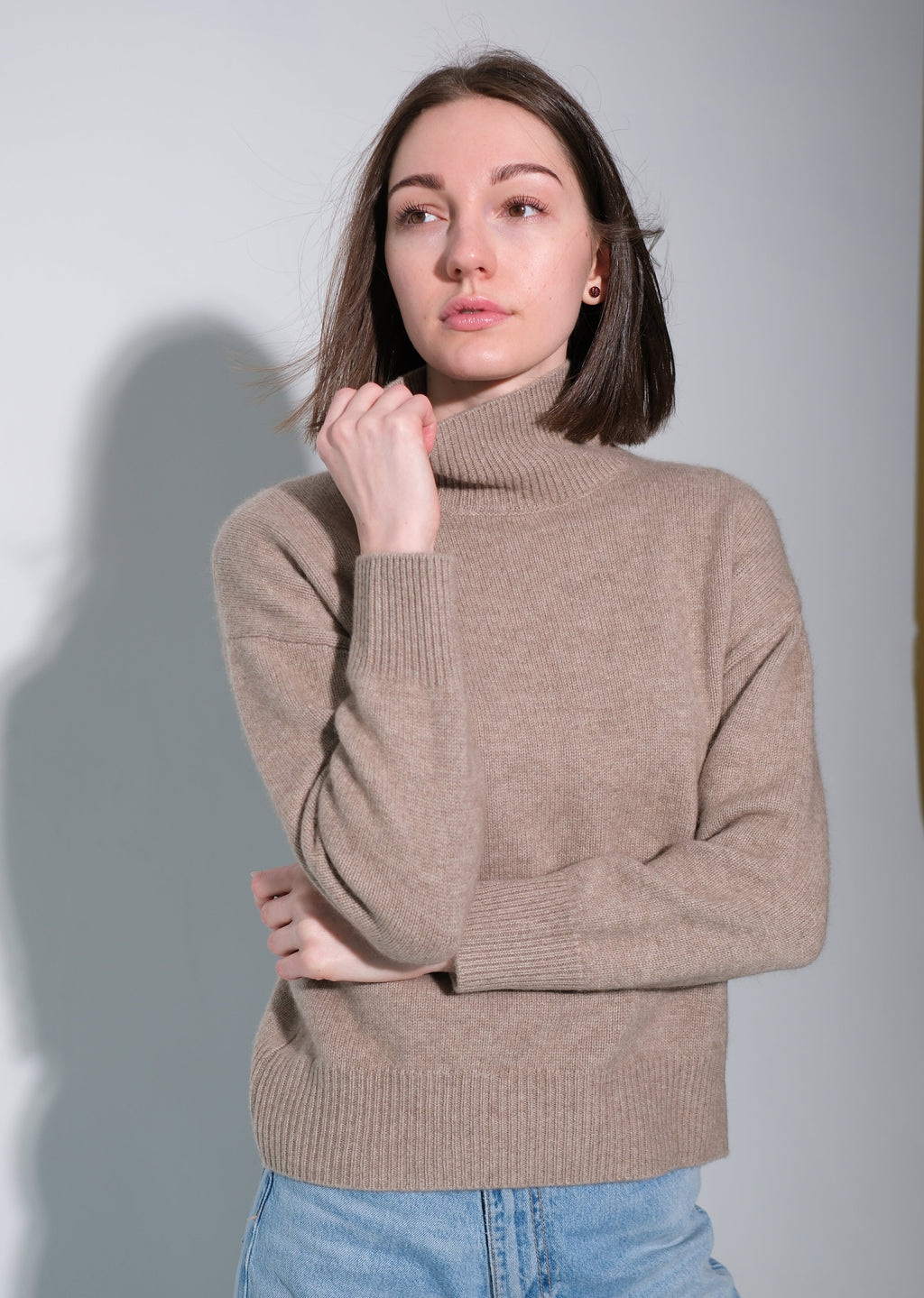 A woman wearing a beige ribbed turtleneck sweater and light blue jeans, standing against a light gray studio background with a soft shadow.