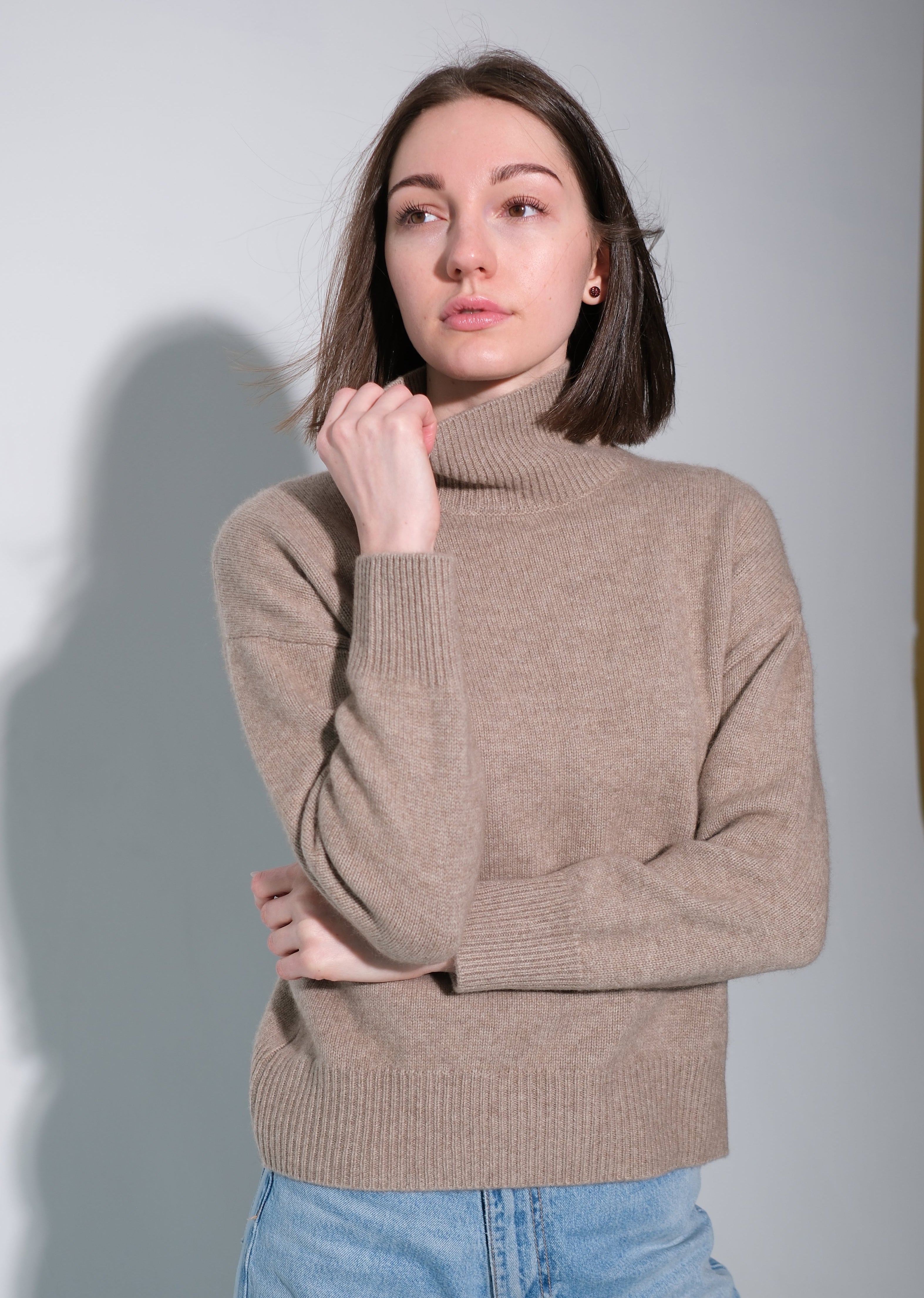 A woman wearing a beige ribbed turtleneck sweater and light blue jeans, standing against a light gray studio background with a soft shadow.