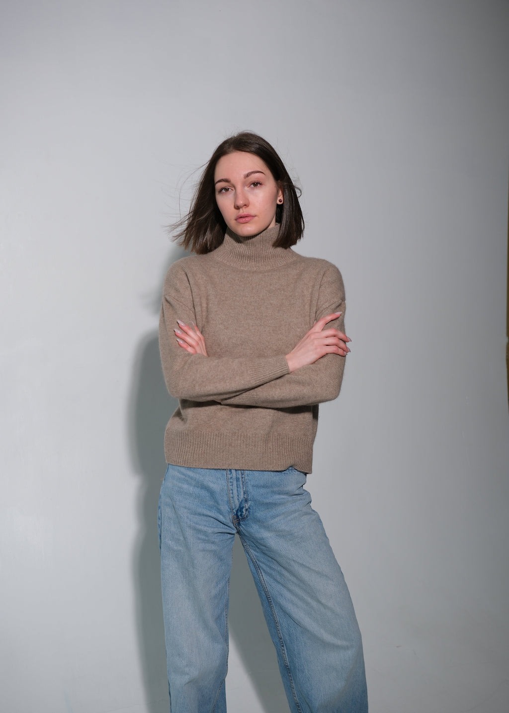 A woman modeling a taupe brown turtleneck sweater and light blue jeans, standing with arms crossed against a plain light gray studio background.