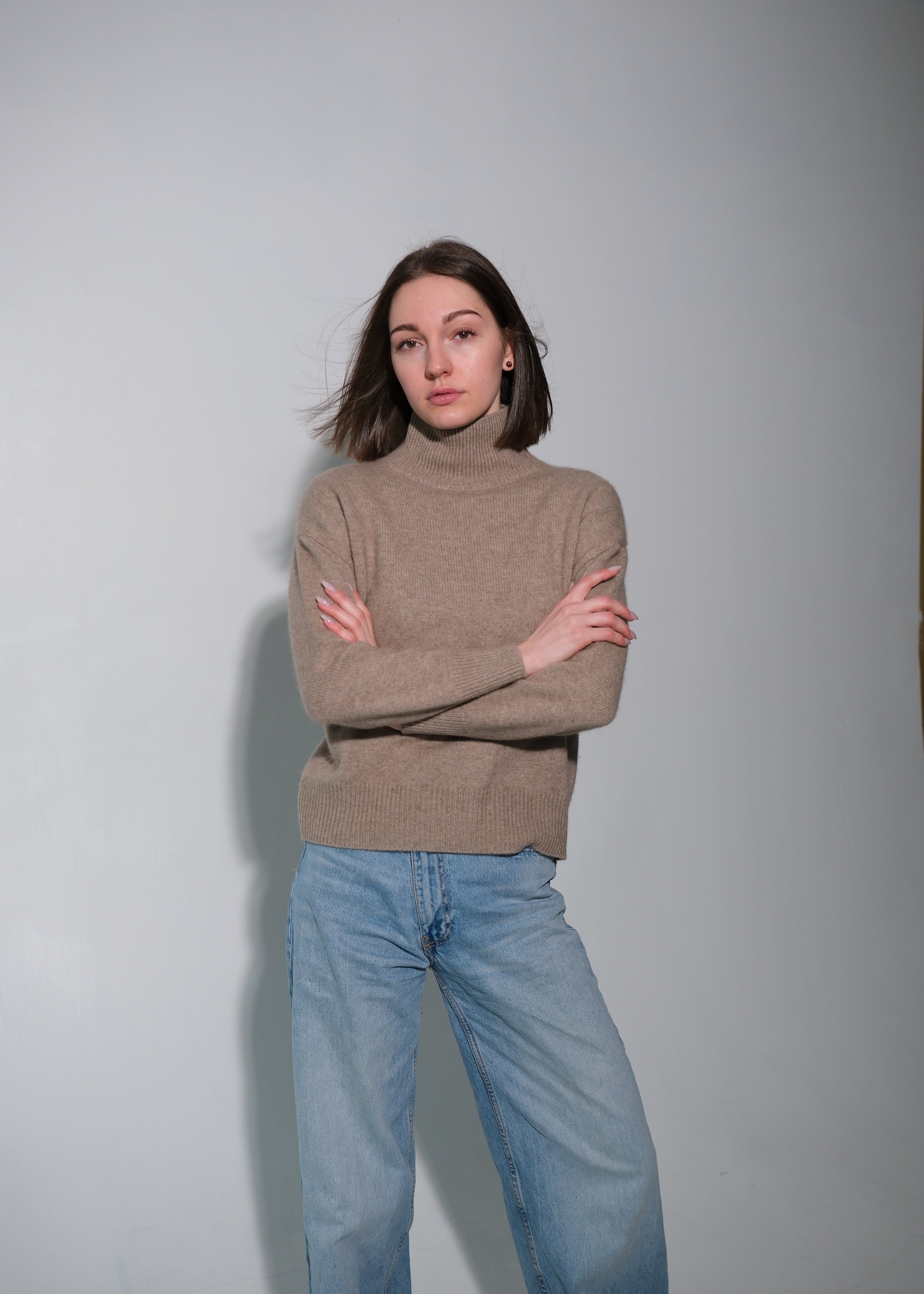 A woman modeling a taupe brown turtleneck sweater and light blue jeans, standing with arms crossed against a plain light gray studio background.