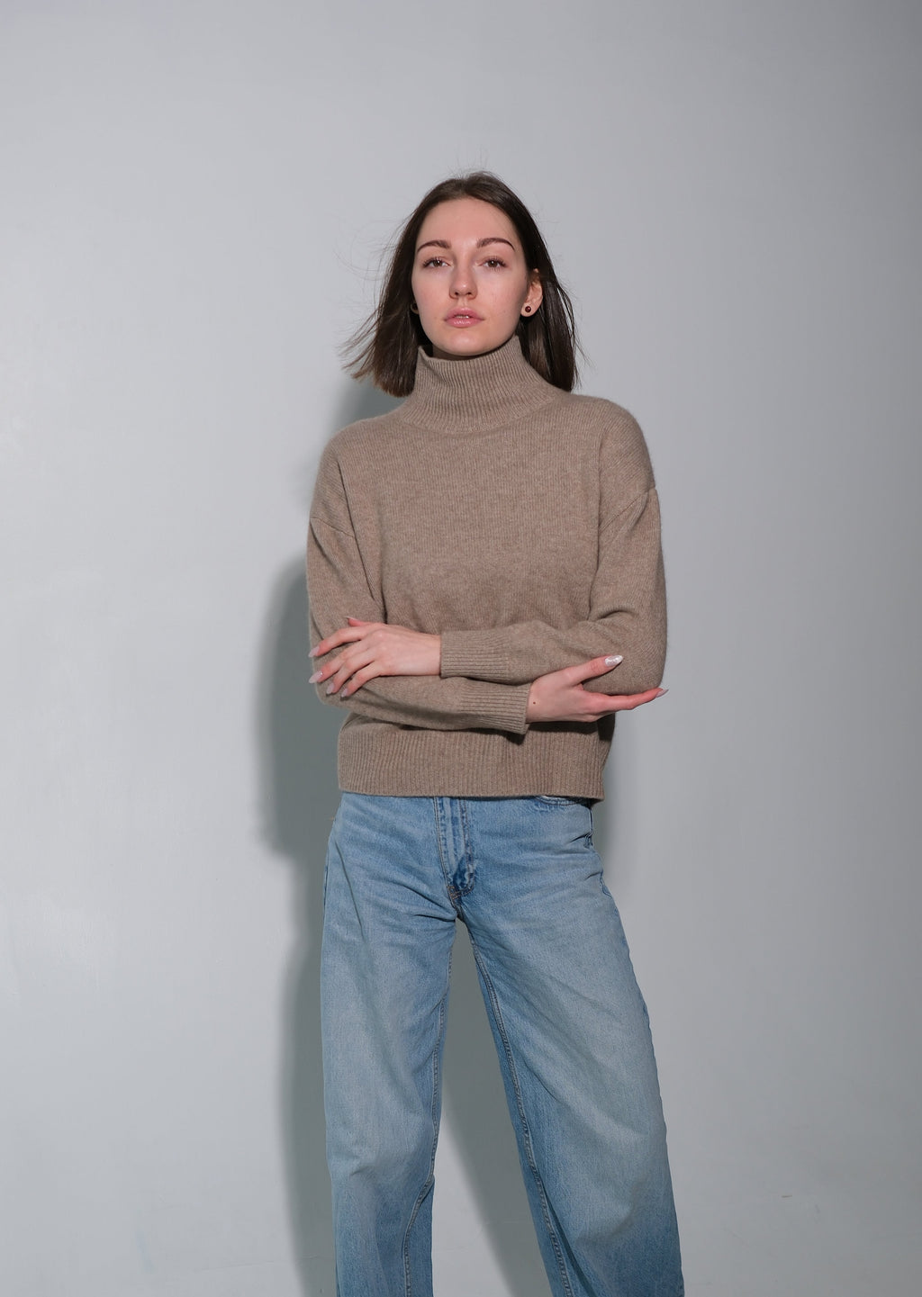 A woman stands against a plain light gray studio background wearing a taupe-brown turtleneck sweater, wide-leg light blue jeans, and gray socks.