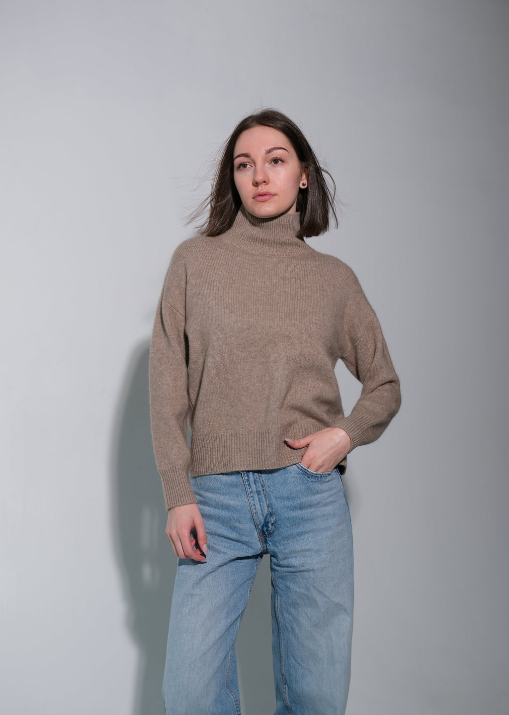 A woman wearing a beige turtleneck sweater and light blue jeans, standing against a plain light gray wall.