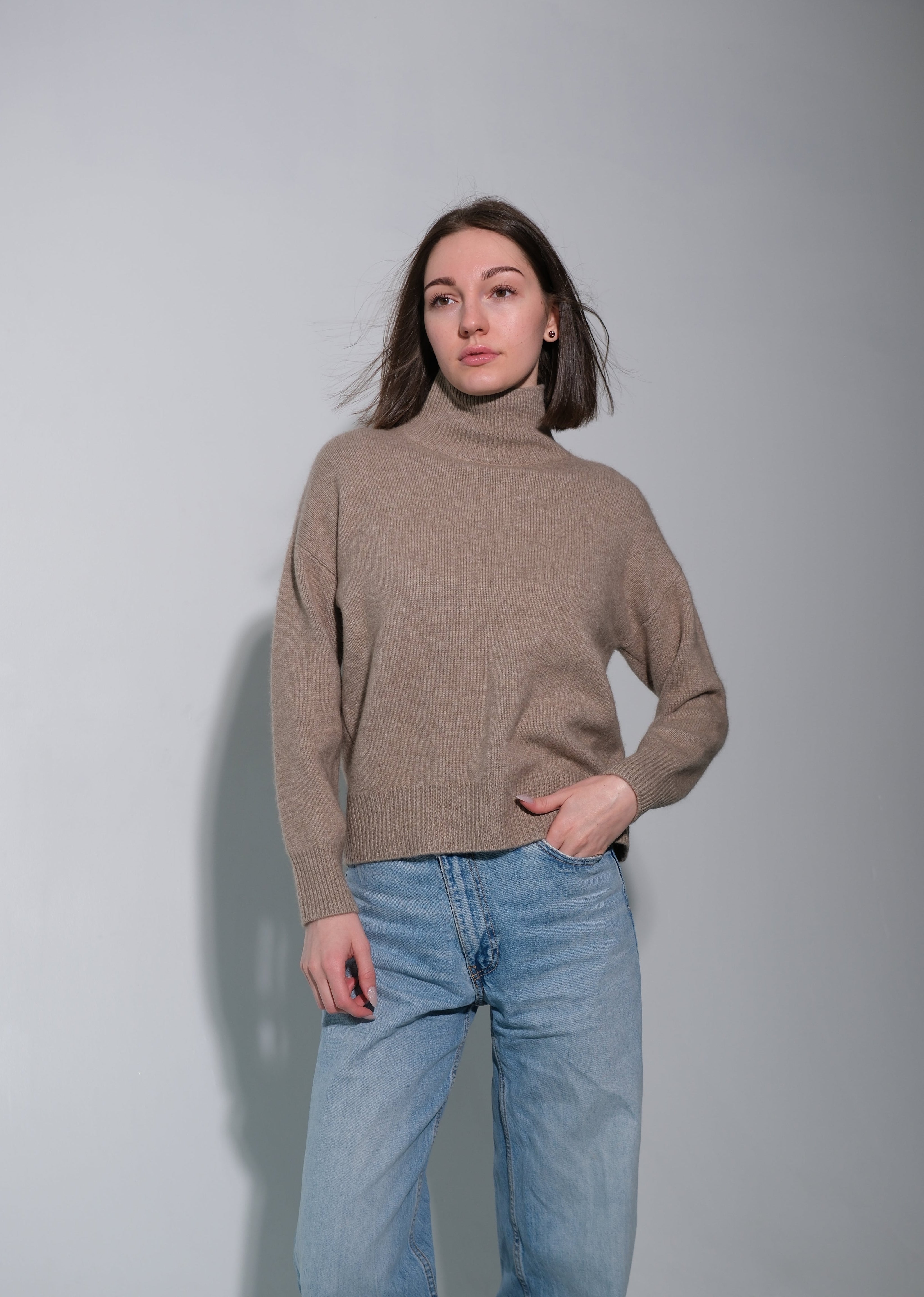 A woman wearing a beige turtleneck sweater and light blue jeans, standing against a plain light gray wall.