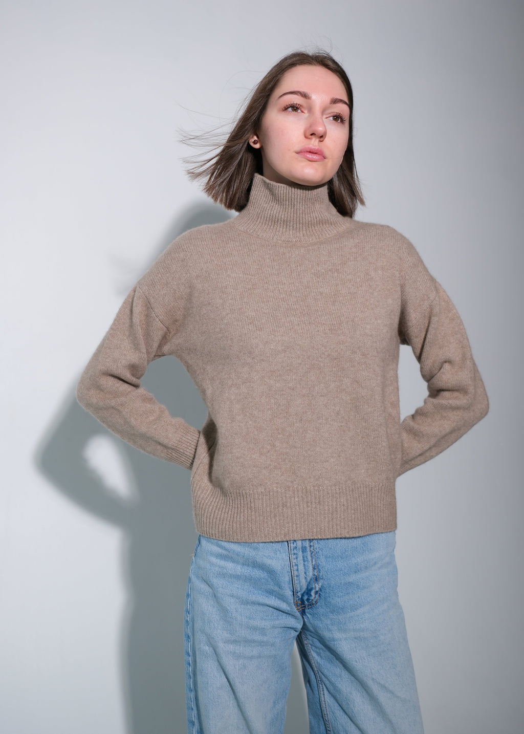 A woman wearing a light brown turtleneck sweater and light blue jeans, standing against a light gray background with her hands on her hips.