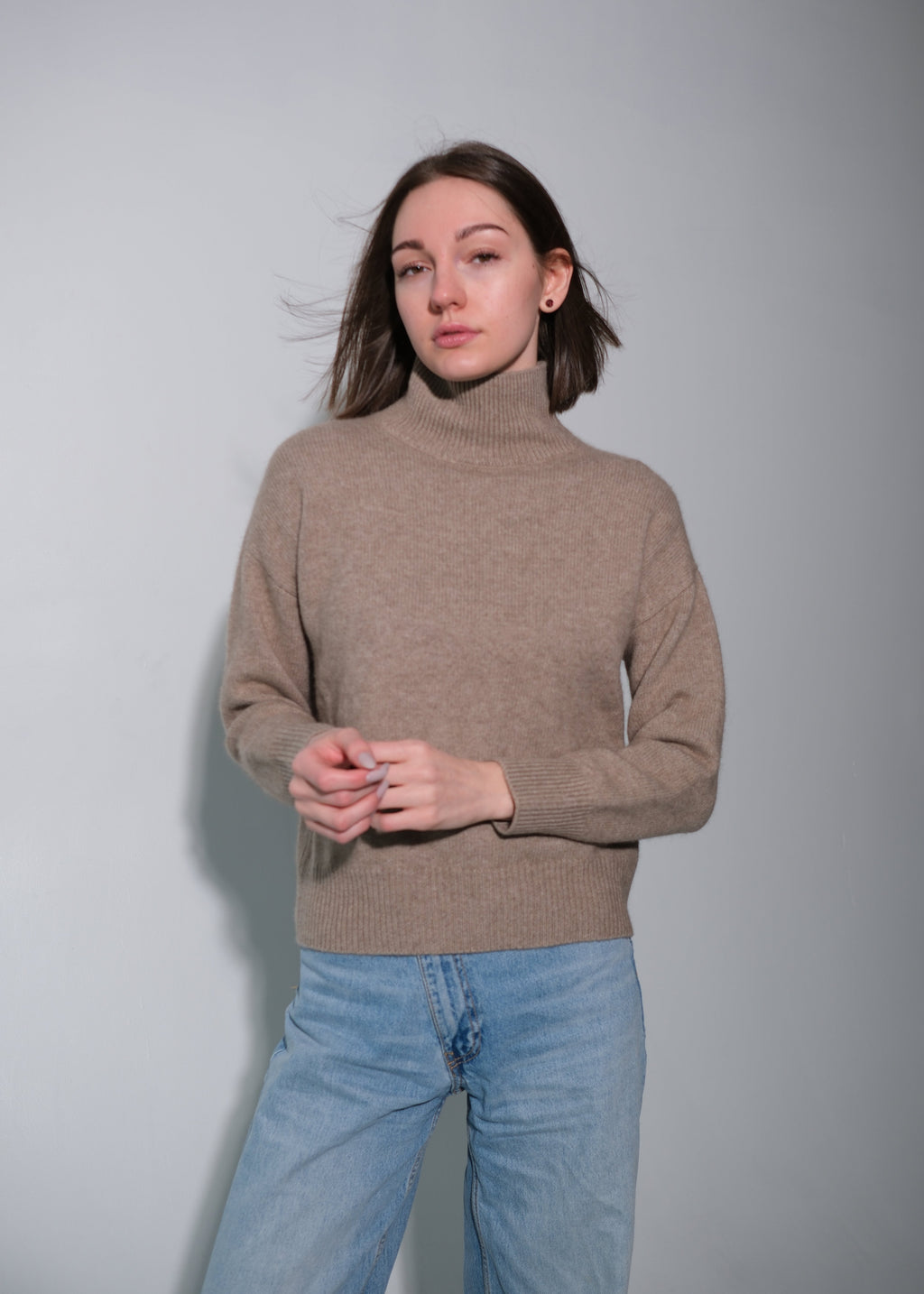 A woman wearing a light brown turtleneck sweater and light blue jeans, standing against a plain gray studio background.