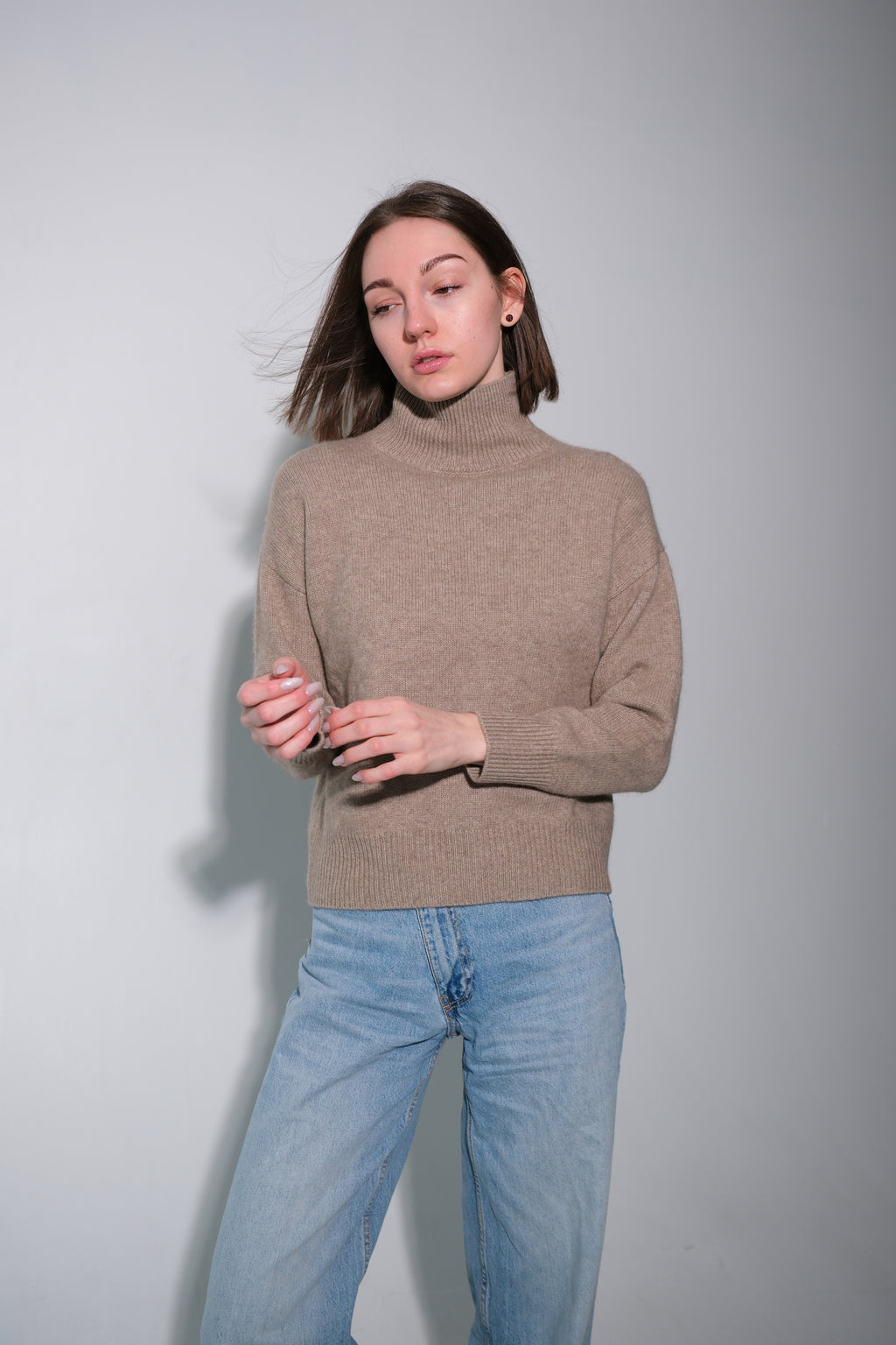 A woman wearing a beige ribbed turtleneck sweater and light blue jeans, standing against a plain light gray studio background.