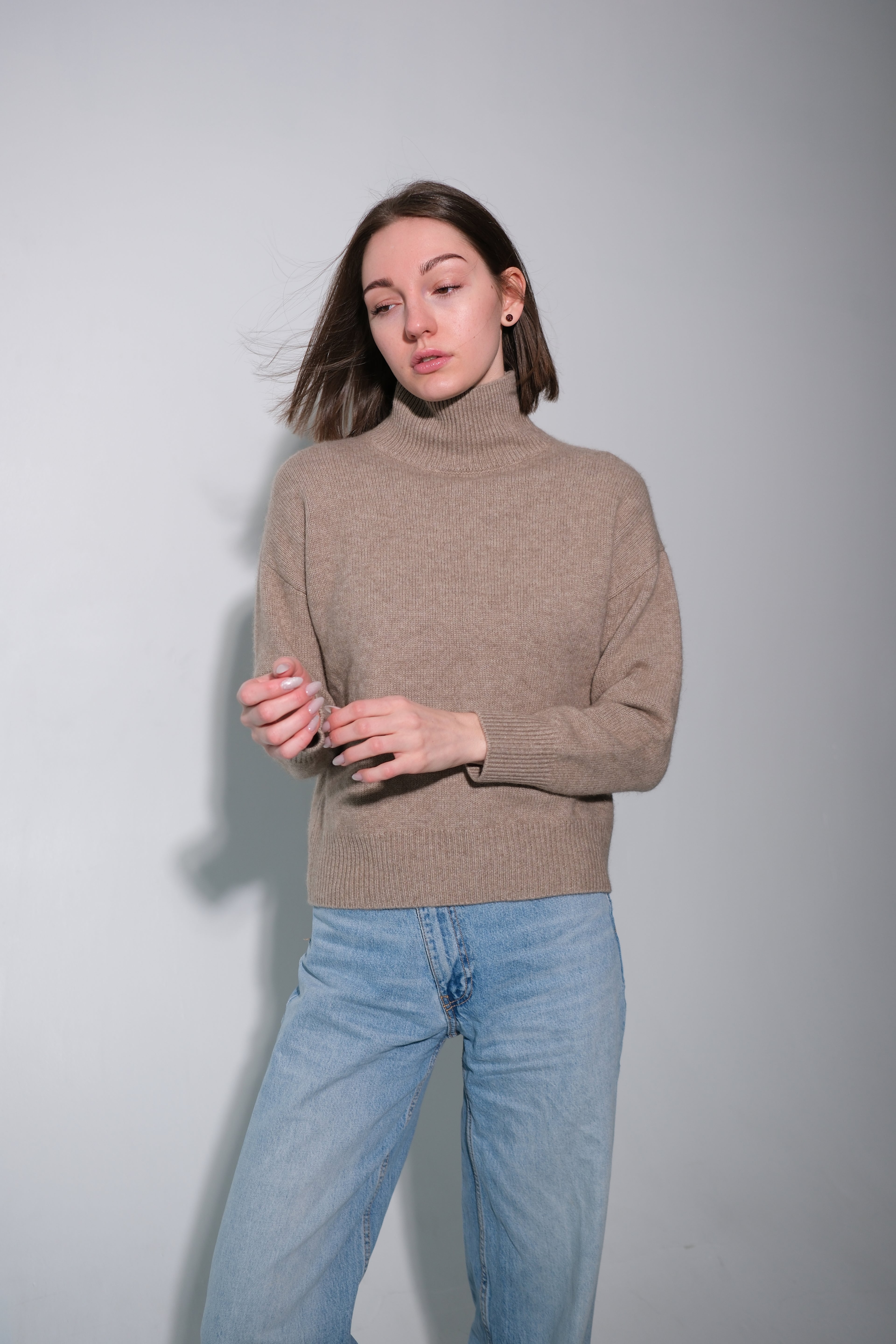A woman wearing a beige ribbed turtleneck sweater and light blue jeans, standing against a plain light gray studio background.