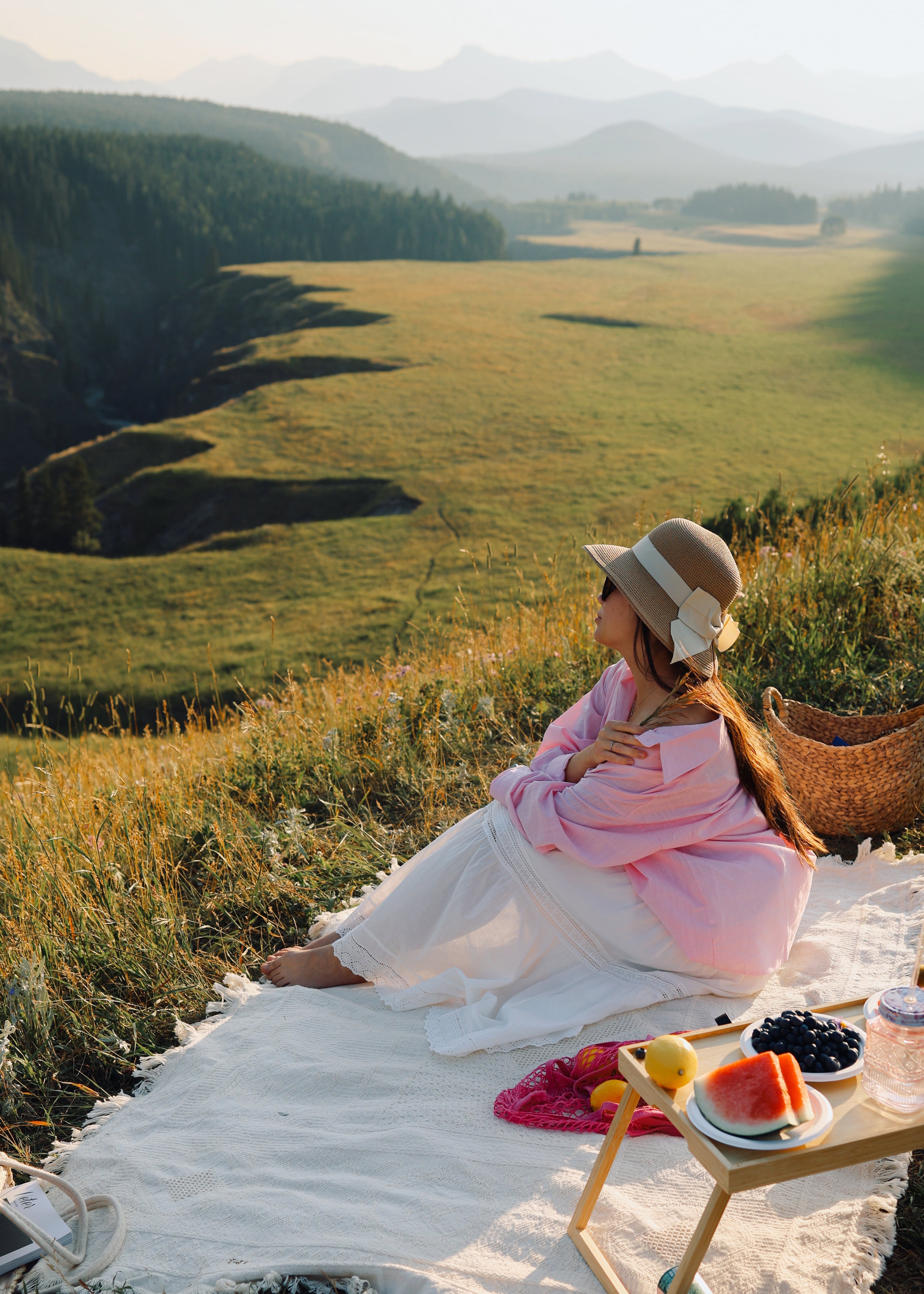 A woman in a pink kimono and straw hat sits on a white blanket in a mountain meadow, with a small picnic table holding fruit and a basket nearby.