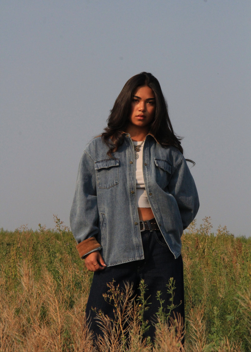 A woman standing in a grassy field wearing an oversized light blue denim jacket over a white crop top and dark pants, photographed outdoors under a pale sky.