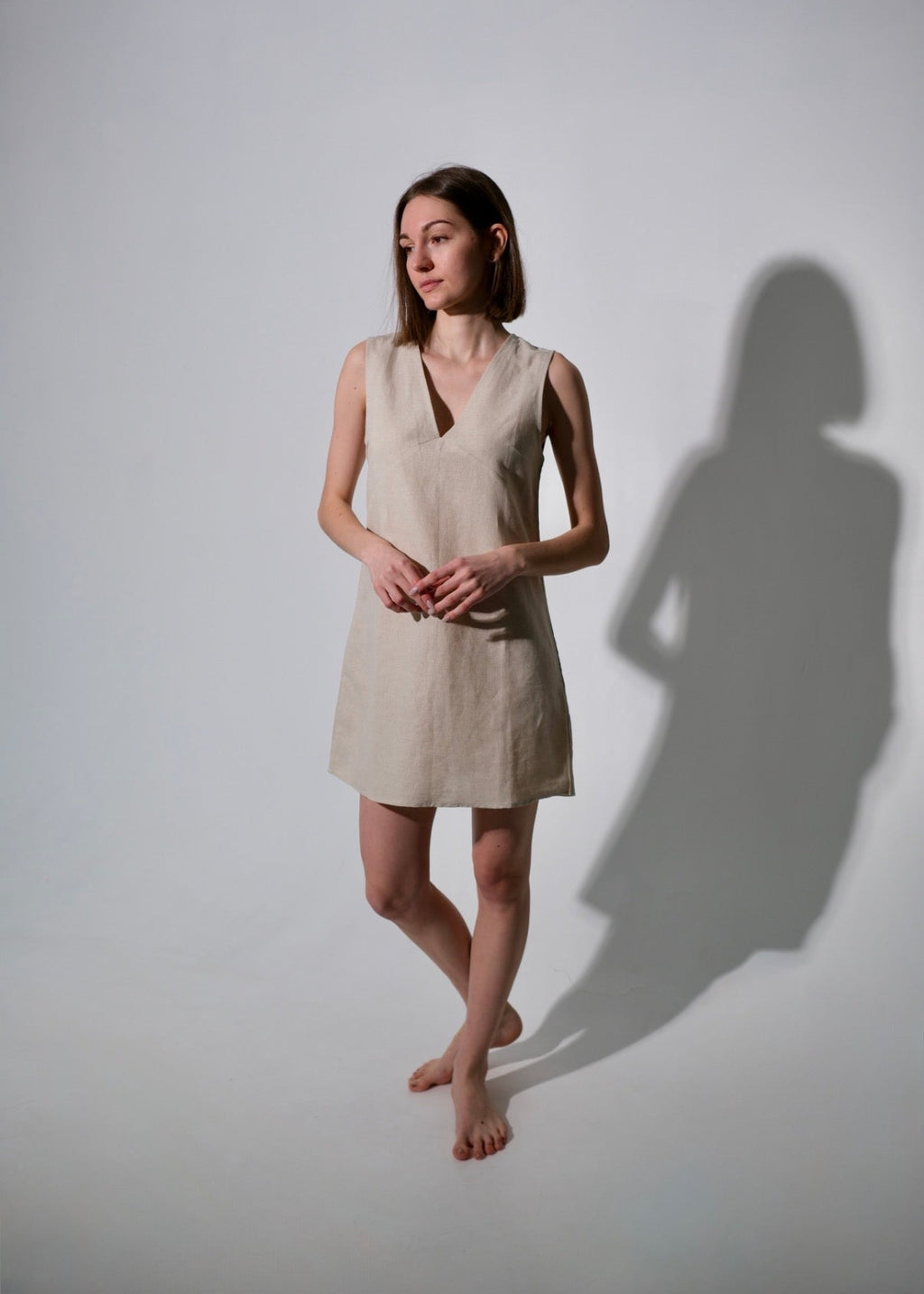 Woman wearing a beige dress standing against a white wall with a soft shadow.