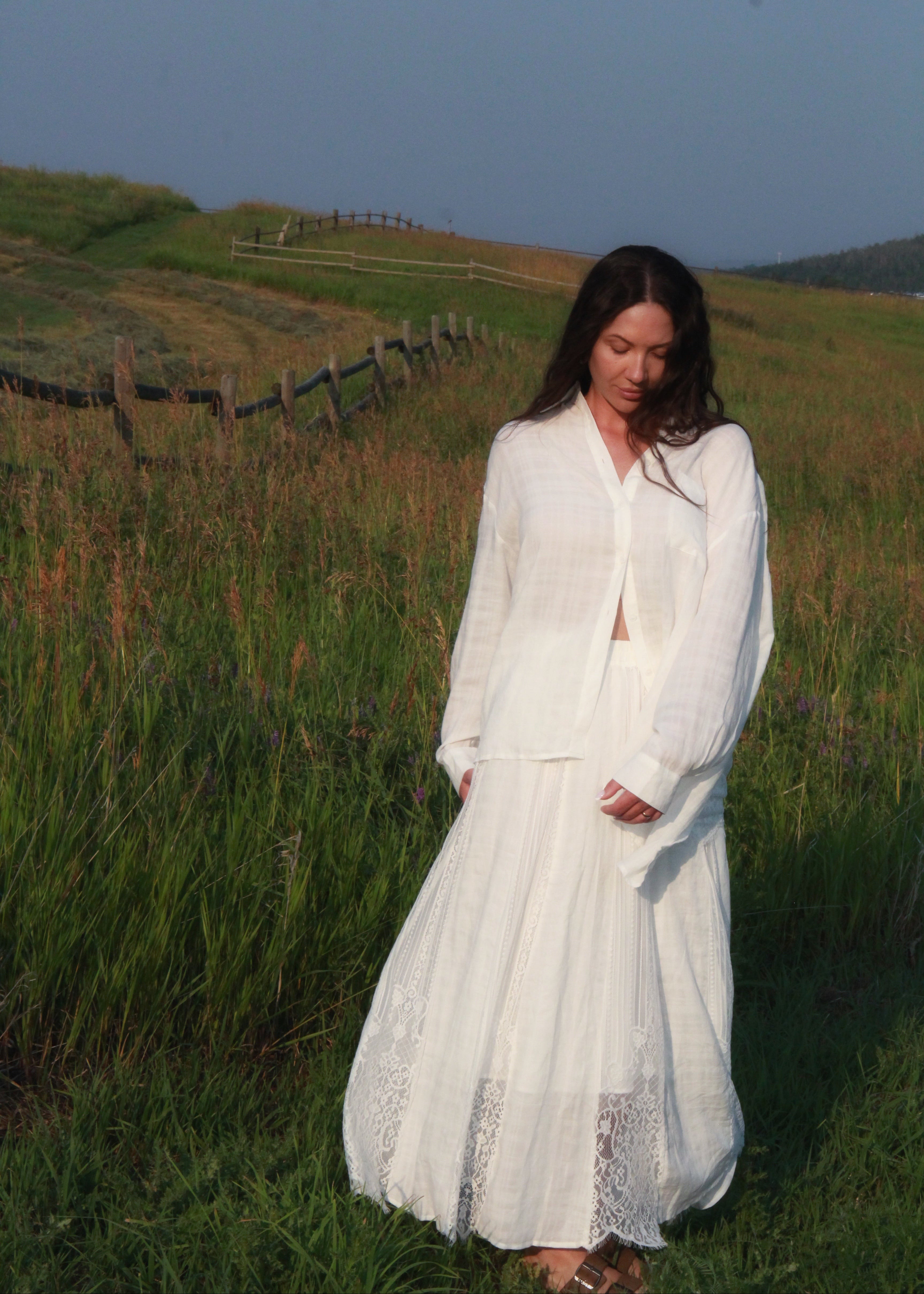 A woman wearing a long-sleeve white button-up shirt and matching white maxi skirt stands in a grassy field with rolling hills in the background.