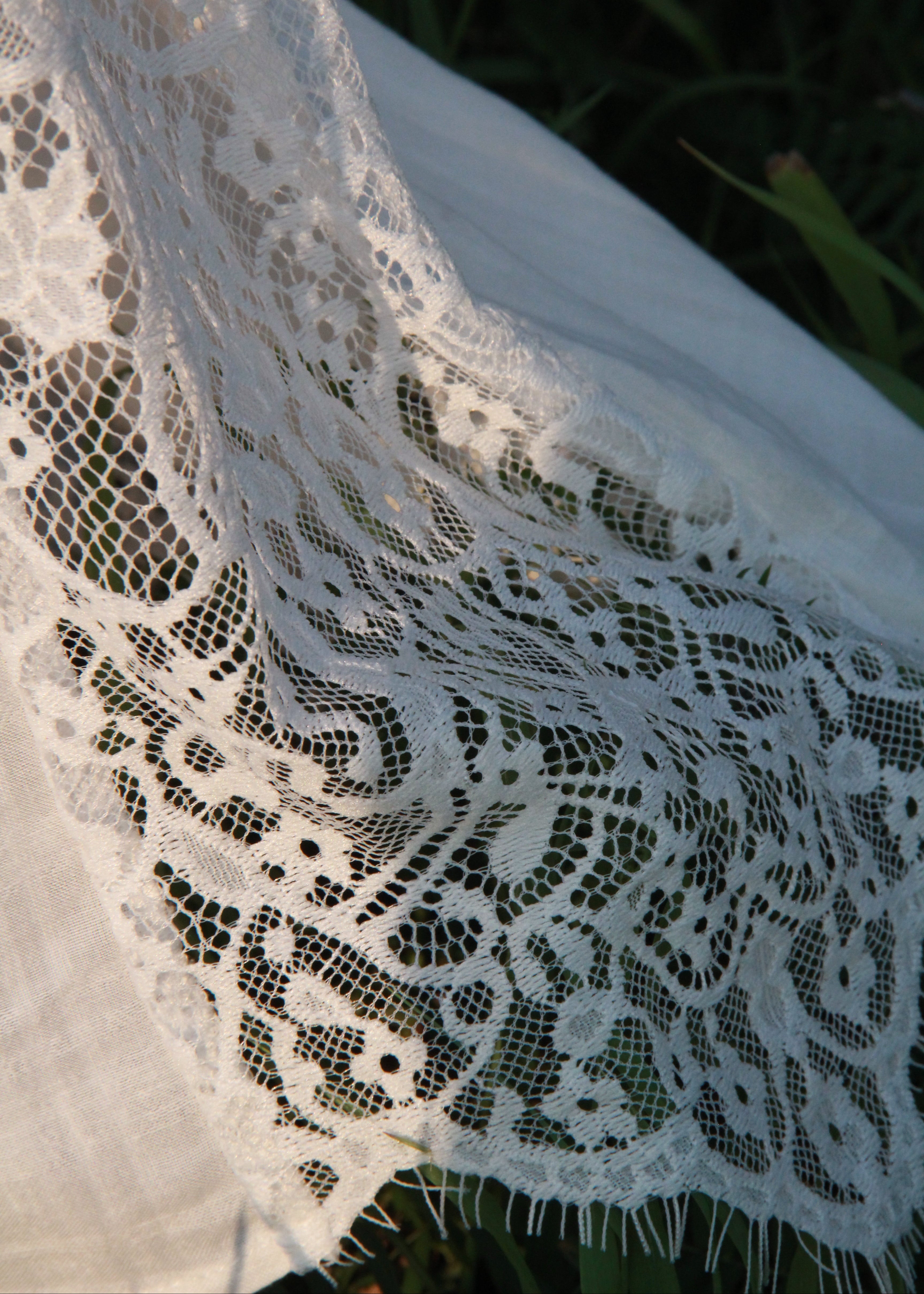 White lace fabric with an intricate floral pattern draped outdoors over dark green grass.