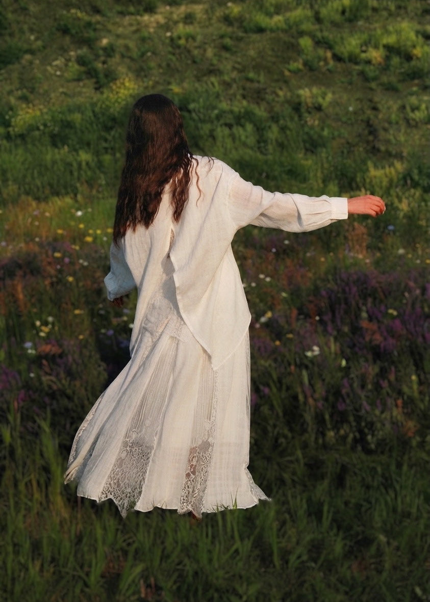 A woman seen from behind wearing a flowing white long-sleeve dress, standing in a grassy outdoor field with her arm extended to the side.