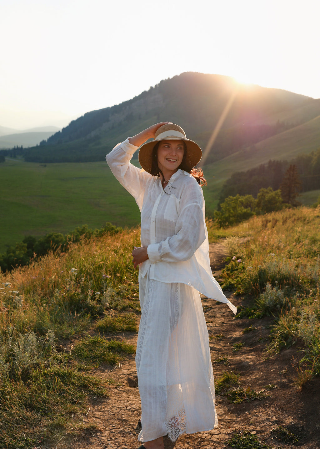 A woman wearing a white long-sleeve button-up shirt and matching long skirt stands on a dirt path in a mountainous field at sunset, holding a wide-brimmed straw hat.