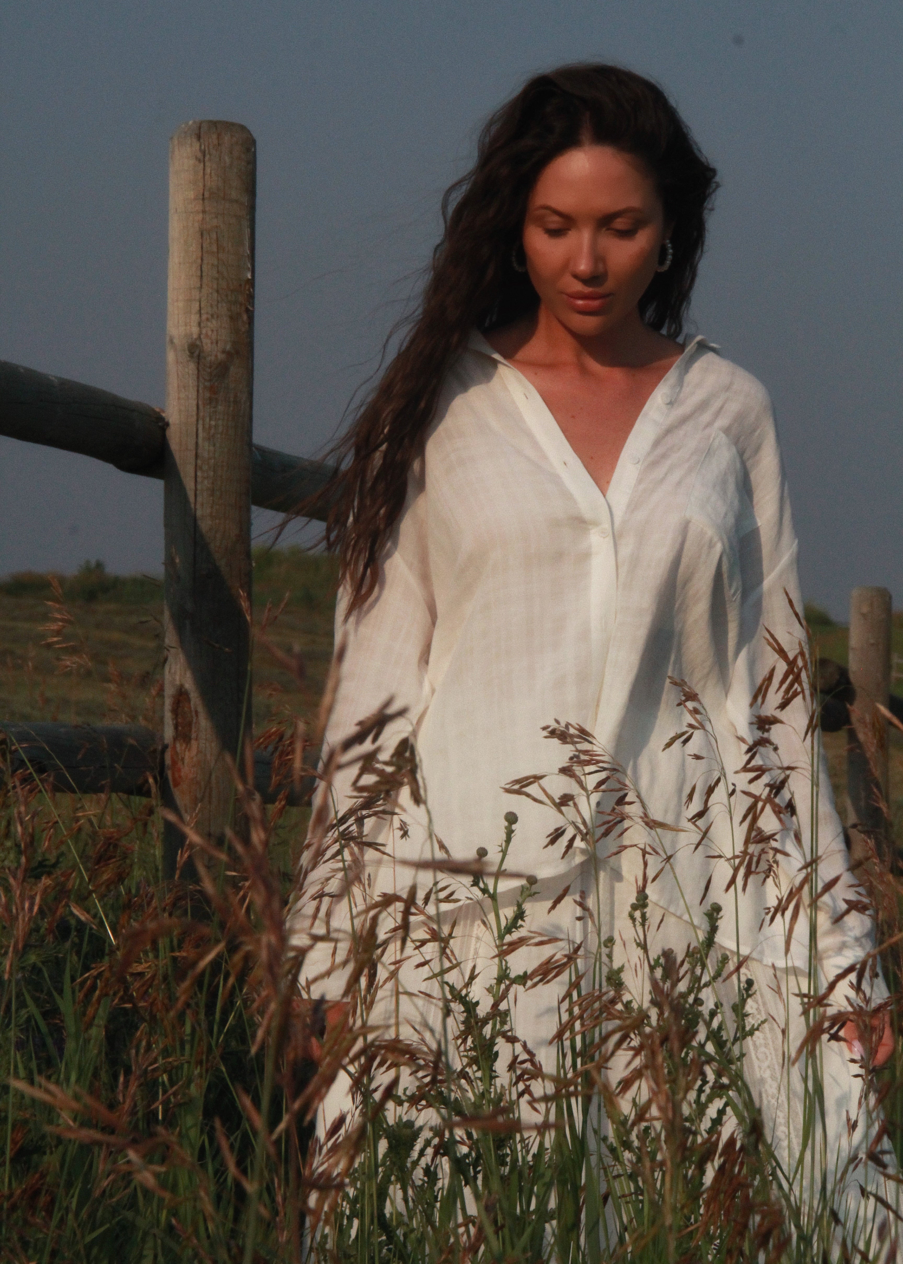 A woman wearing a loose white long-sleeve dress standing outdoors in tall grass near a wooden fence under a blue sky.