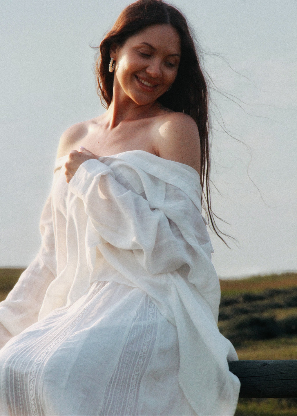 A woman with long dark hair wearing a flowing white off-the-shoulder dress, sitting outdoors against a pale sky with a blurred field in the background.
