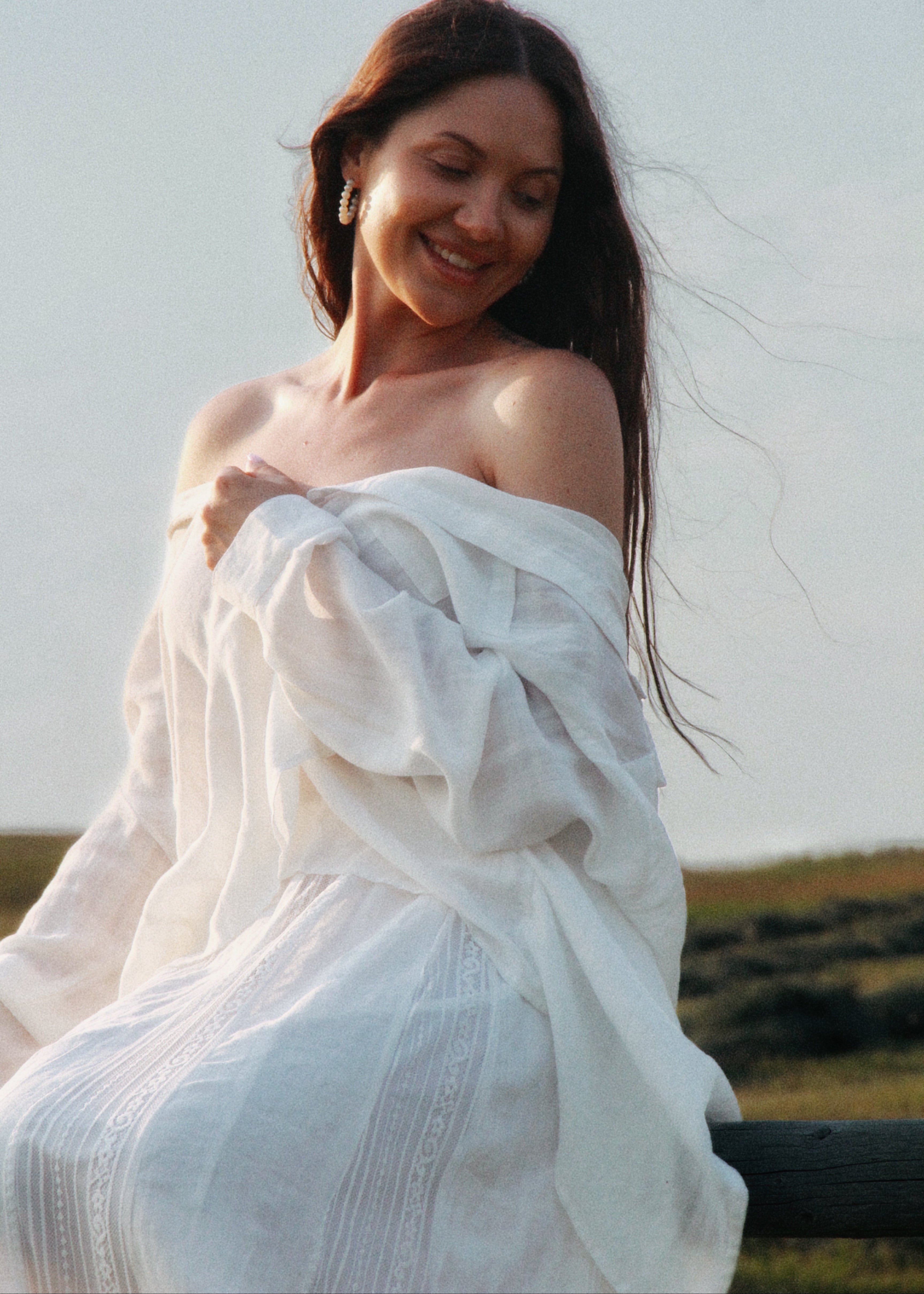 A woman with long dark hair wearing a flowing white off-the-shoulder dress, sitting outdoors against a pale sky with a blurred field in the background.
