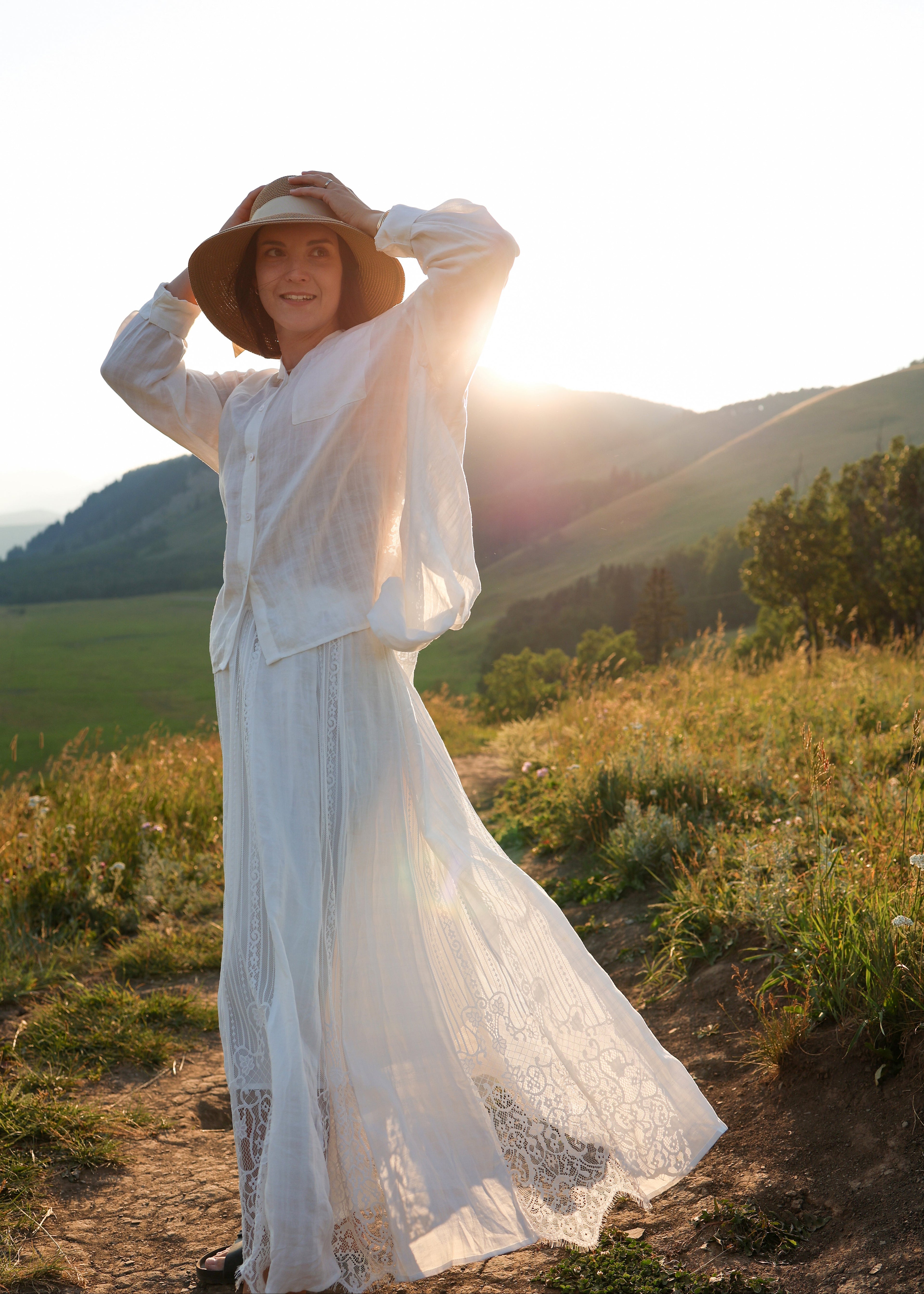 A woman wearing a long white lace dress and a wide-brimmed straw hat stands outdoors on a grassy hillside at sunset, with mountains in the background.