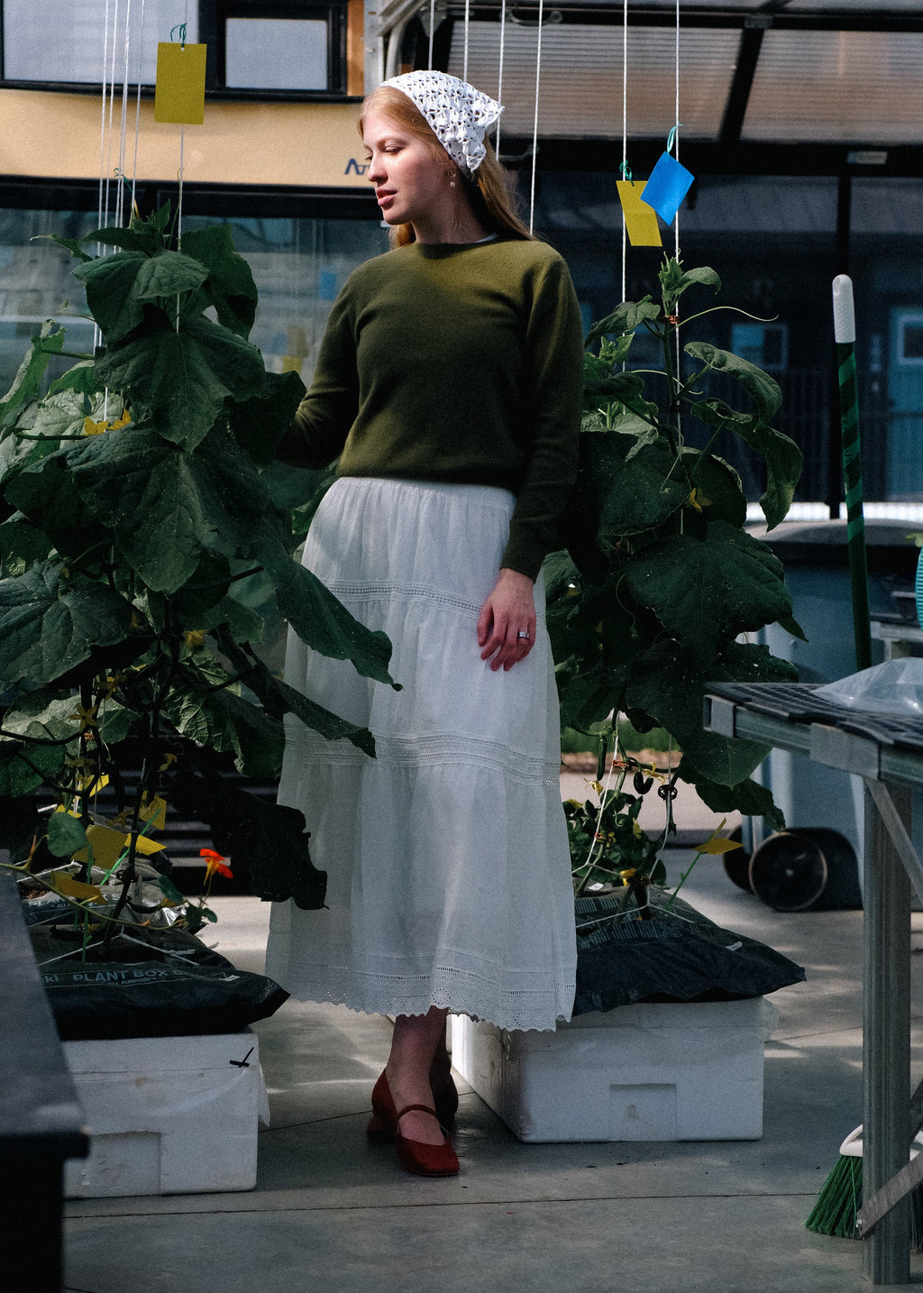 Woman in a green sweater and white skirt standing in a greenhouse with plants and tables around her.