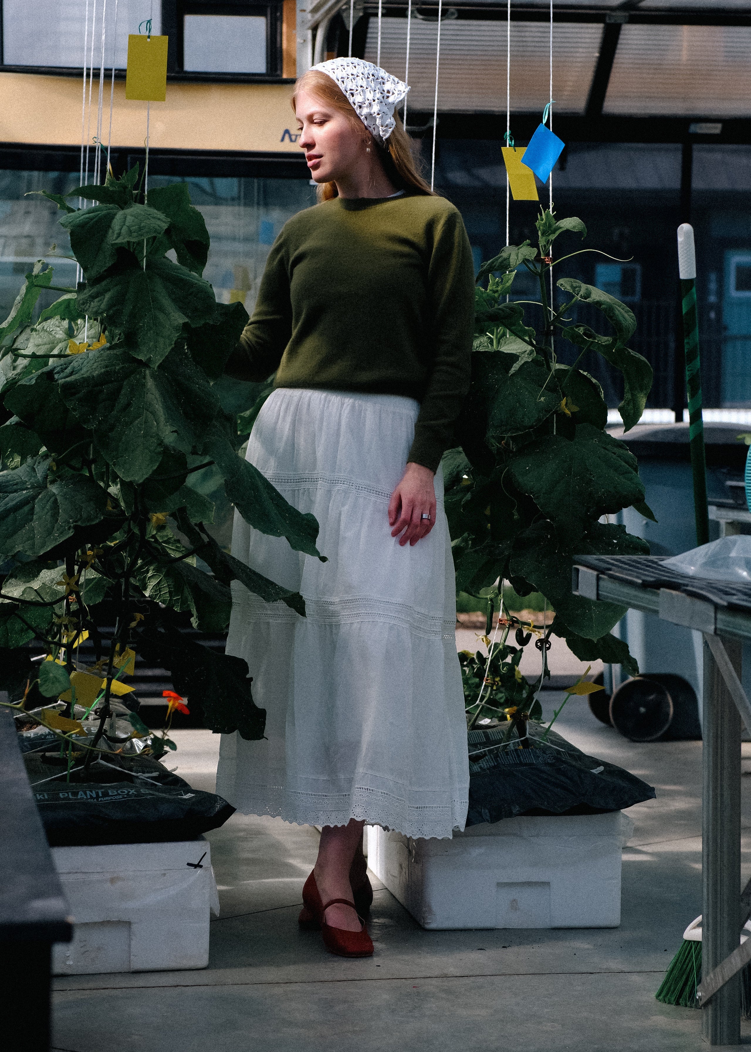 Woman in a green sweater and white skirt standing in a greenhouse with plants and tables around her.