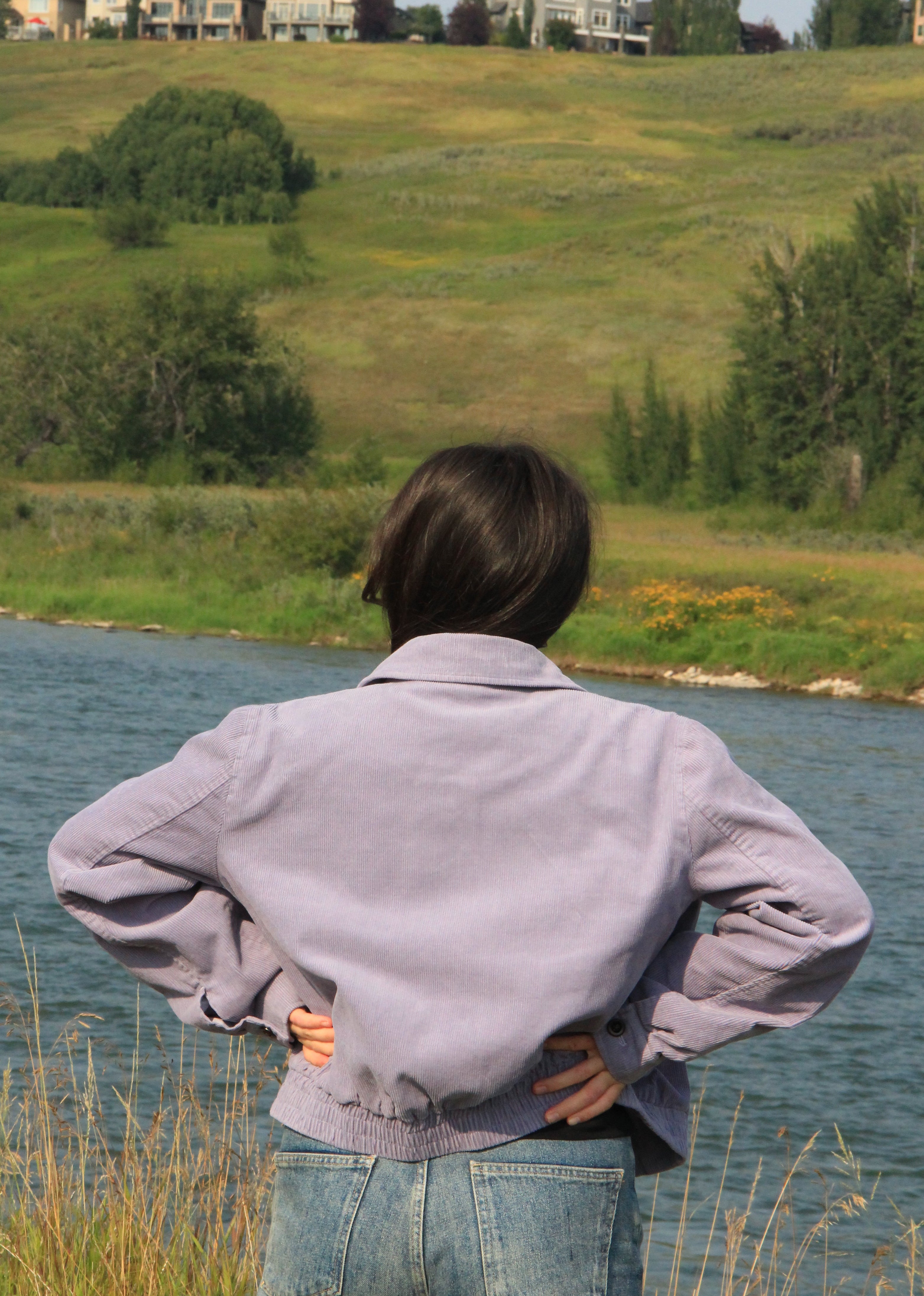 A person seen from behind wearing a light purple-gray button-up shirt and blue jeans, standing in tall grass by a river and looking toward a grassy hill with houses in the distance.