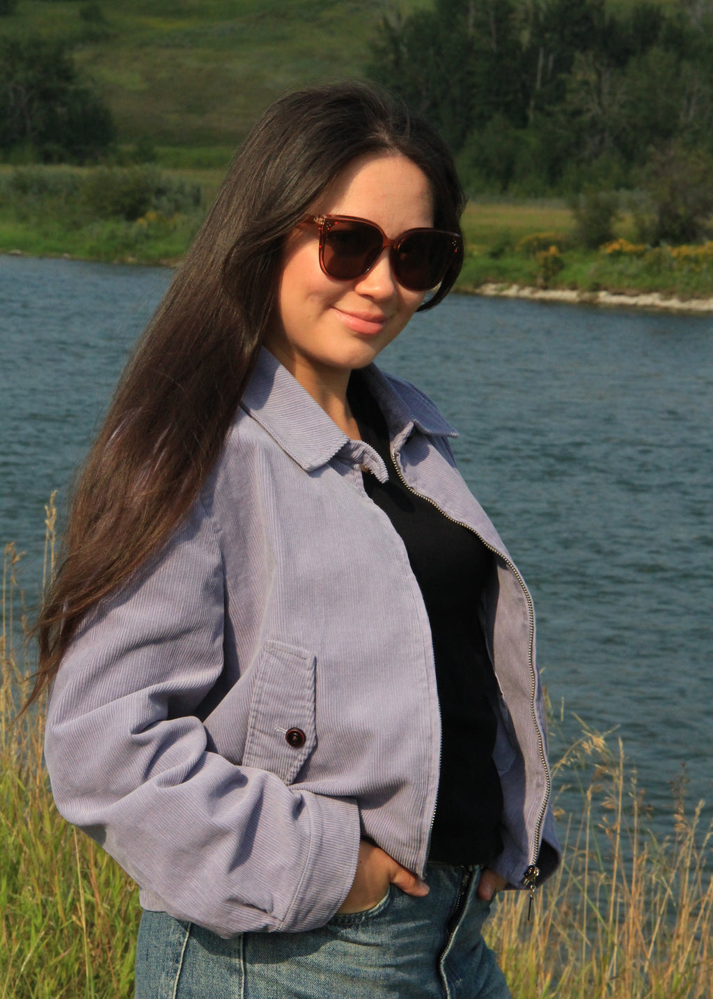 A woman with long dark hair wearing a light purple corduroy jacket over a black top and blue jeans, standing outdoors near a river with grass and trees in the background.